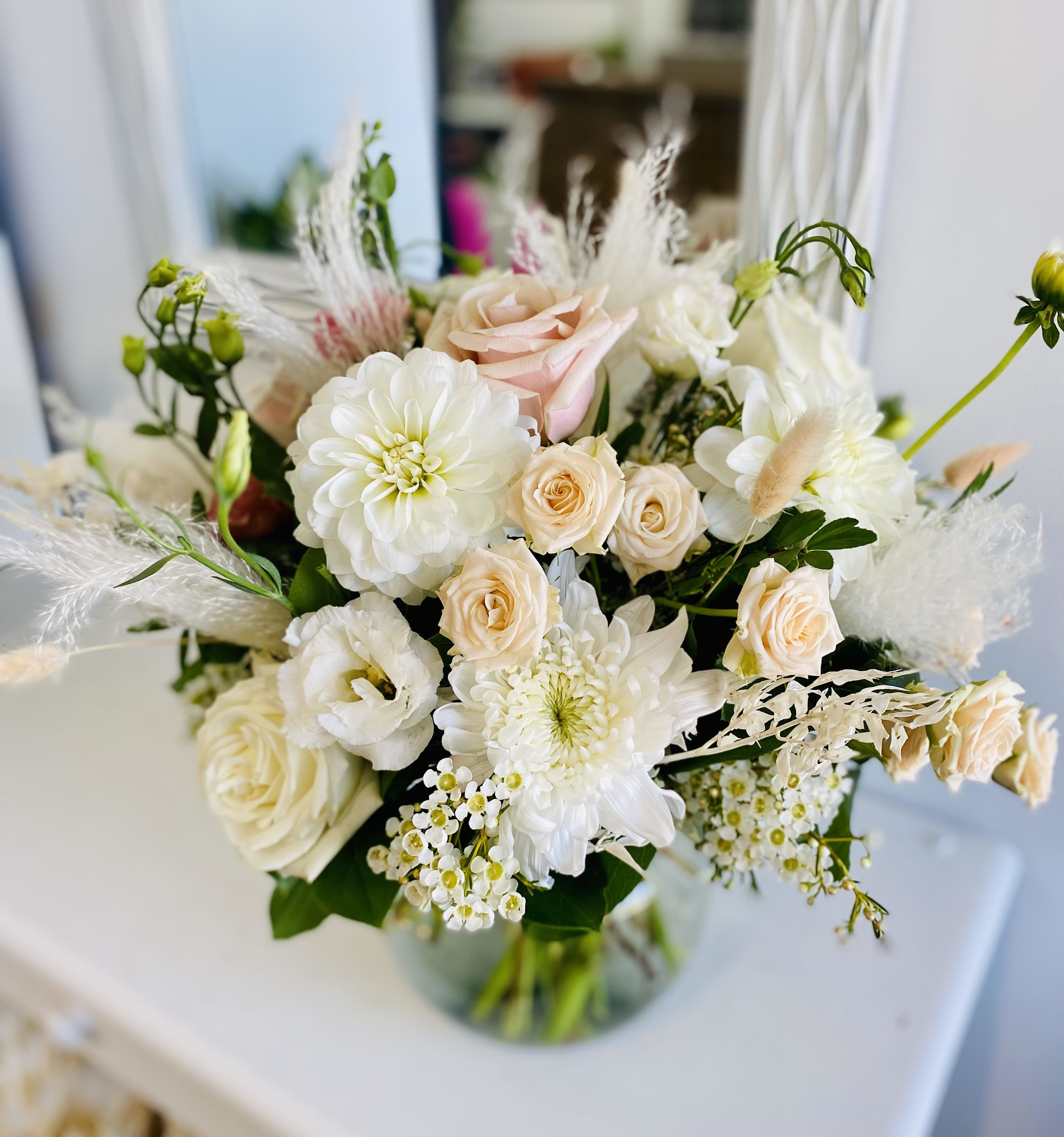 Elegant Blooms Elegant Blooms - Elegant and ethereal, this soft-toned arrangement exudes timeless sophistication. Creamy white dahlias, blush roses, lisianthus, and fluffy mums are beautifully accented with delicate waxflower, airy pampas grass, and soft greenery. Arranged in a round glass vase, it’s the perfect piece for weddings, sympathy, or simply adding a graceful touch to any setting.