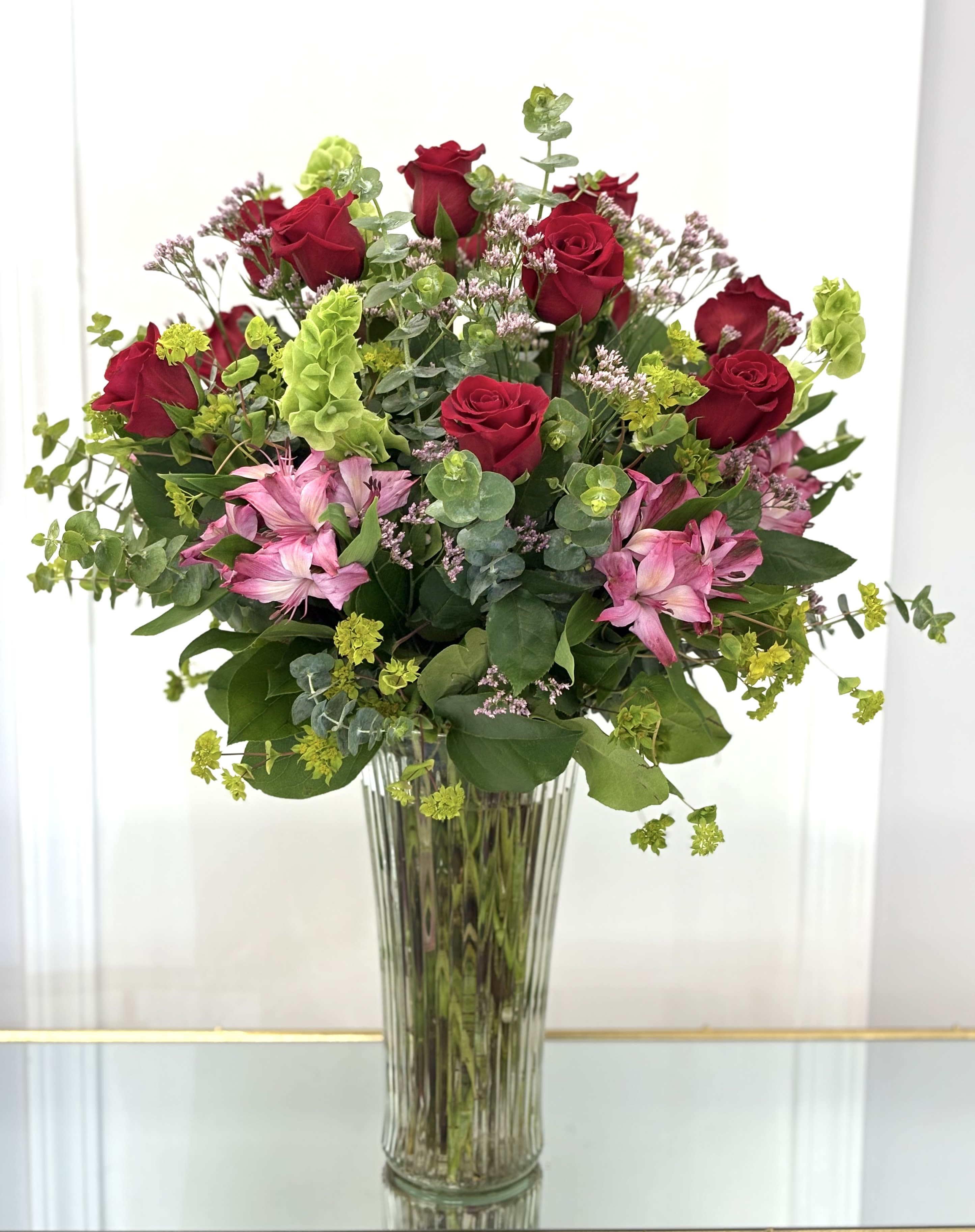 Crimson Meadow By VLM Flowers - A graceful celebration of romance and vitality, Crimson Meadow combines classic red roses with cheerful pink alstroemeria and playful sprigs of green bells of Ireland. Hints of eucalyptus and delicate filler blooms complete the look, adding texture and fragrance. Designed in a tall fluted vase, this arrangement is perfect for anniversaries, heartfelt congratulations, or simply to brighten someone’s day with elegance and warmth.