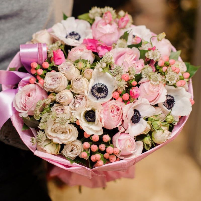 Pink Secret - This breathtaking arrangement is a bold yet refined medley of textures and hues, perfect for making a statement. Light purple spray roses introduce a soft, dreamy elegance, while vibrant hot pink mini carnations infuse the bouquet with playful energy and depth.  The lush pink hypericum berries add an unexpected pop of texture, complementing the romantic allure of the large pink roses, whose full, velvety petals captivate with their timeless beauty. Delicate white astrantia blooms contribute an ethereal charm, their intricate starburst-like forms adding an air of sophistication. Completing the ensemble, the pristine white anemones, with their striking dark centers, create a striking contrast that ties the arrangement together with contemporary flair.  Ideal for celebrating love, joy, or any special moment, this arrangement combines vivid pops of color with subtle accents to create a dynamic and unforgettable presentation. Whether it’s for a joyful celebration or a heartfelt gesture, this bouquet captures the essence of beauty, passion, and grace in every petal.