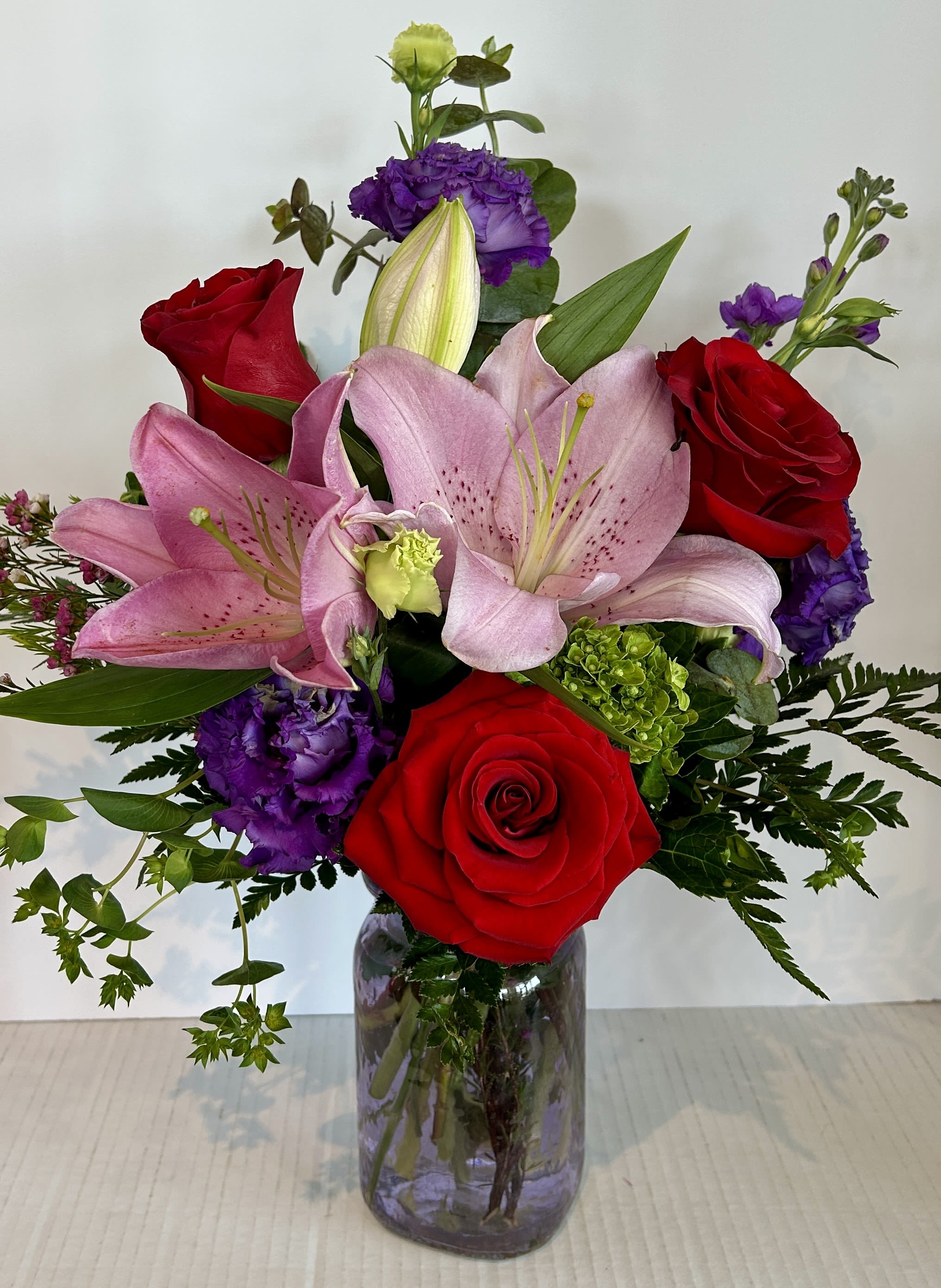 Hat's Off to You - A lavish bouquet of cherished and fragrant flowers featuring lilies, roses, stock, and lisianthus. This arrangement is designed in a purple vase and is styled with elegant greens, wax flowers, and a green hydrangea.     (Substitutions may be required based on availability) 