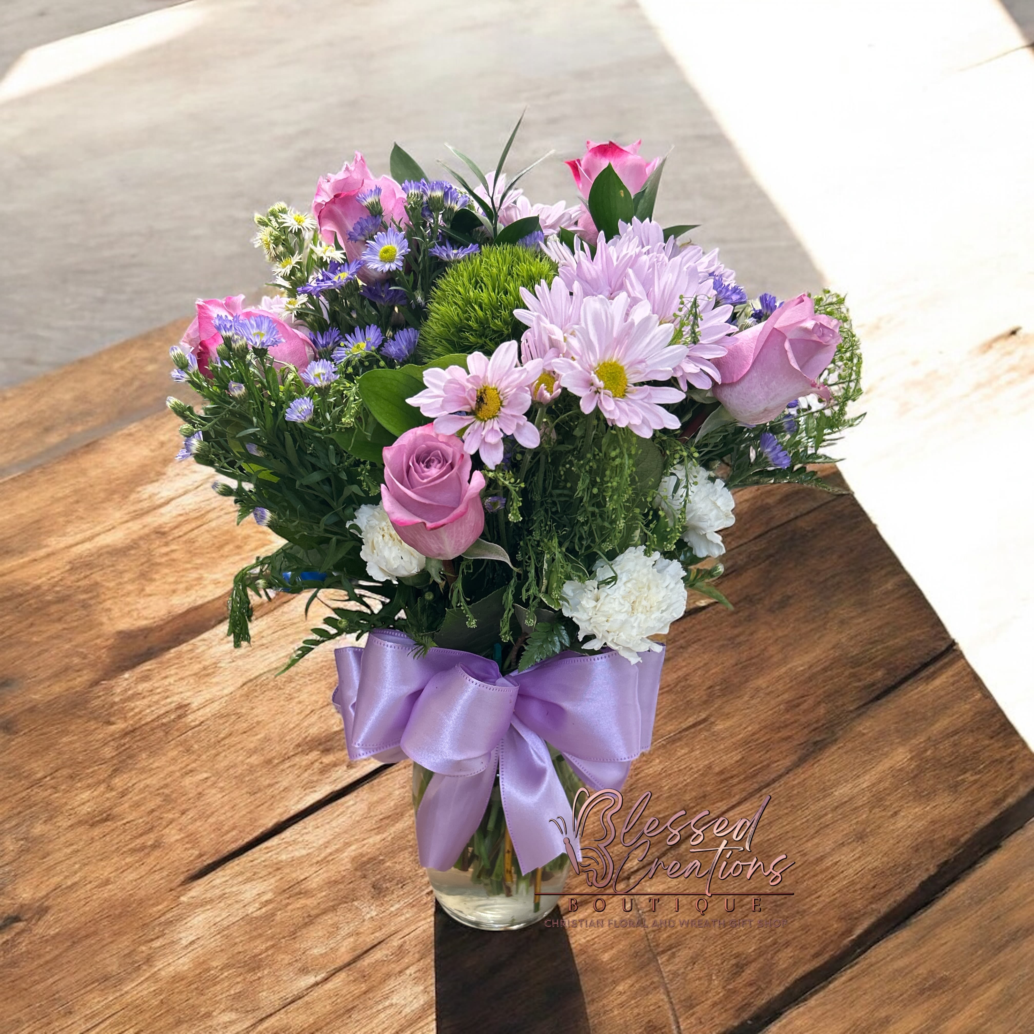 Faithful Elegance  - A stunning arrangement of purple roses, lavender daisies, and white cushion mums, gracefully accented with lush greenery and filler blooms. Presented in a clear vase tied with a soft purple bow, this design radiates peace, hope, and the gentle beauty of God’s promises. Perfect for sending love, encouragement, or a reminder of His faithfulness.