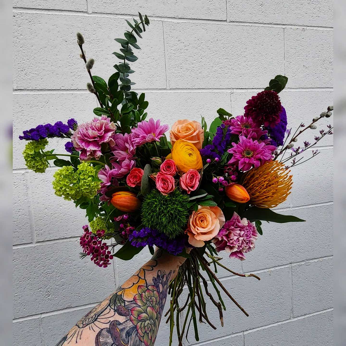 Spring Lux Bouquet Spring Lux Bouquet - A vibrant blend of fresh spring blooms in shades of orange, yellow, purple, mauve, and green designed with lush mixed greenery into a hand-tied cut bouquet.