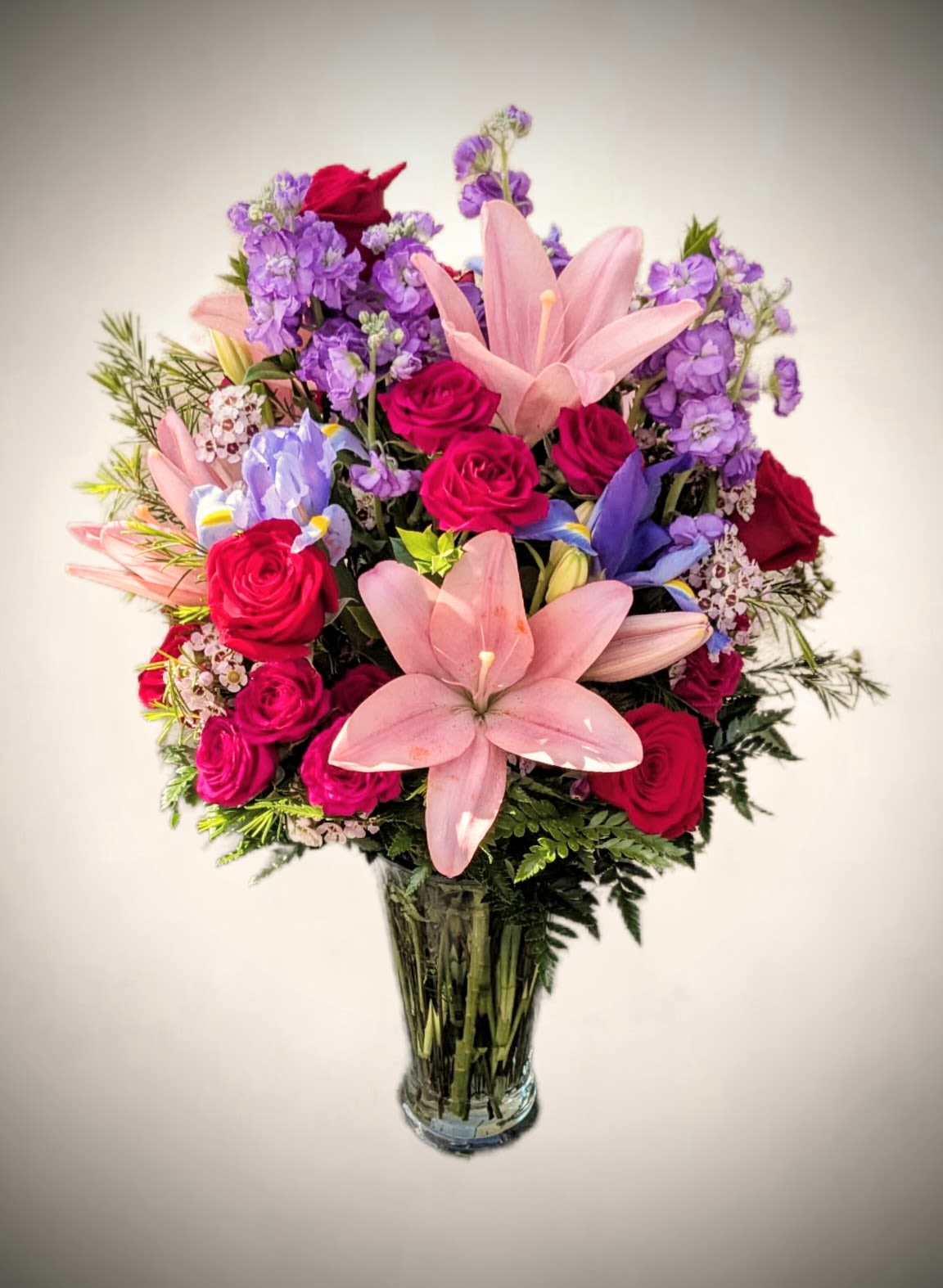 Radiance Romance Vase Arrangement Radiance Romance Vase Arrangement - Bright, bold, and bursting with beauty, this stunning arrangement is a showstopper for any occasion. Featuring a vibrant mix of hot pink roses, soft pink lilies, delicate purple stock, blue iris, and touches of waxflower and lush greenery, this bouquet brings a perfect balance of elegance and cheer. Expertly arranged in a classic clear glass vase, it’s ideal for birthdays, anniversaries, or simply to brighten someone’s day with a splash of color and love.
