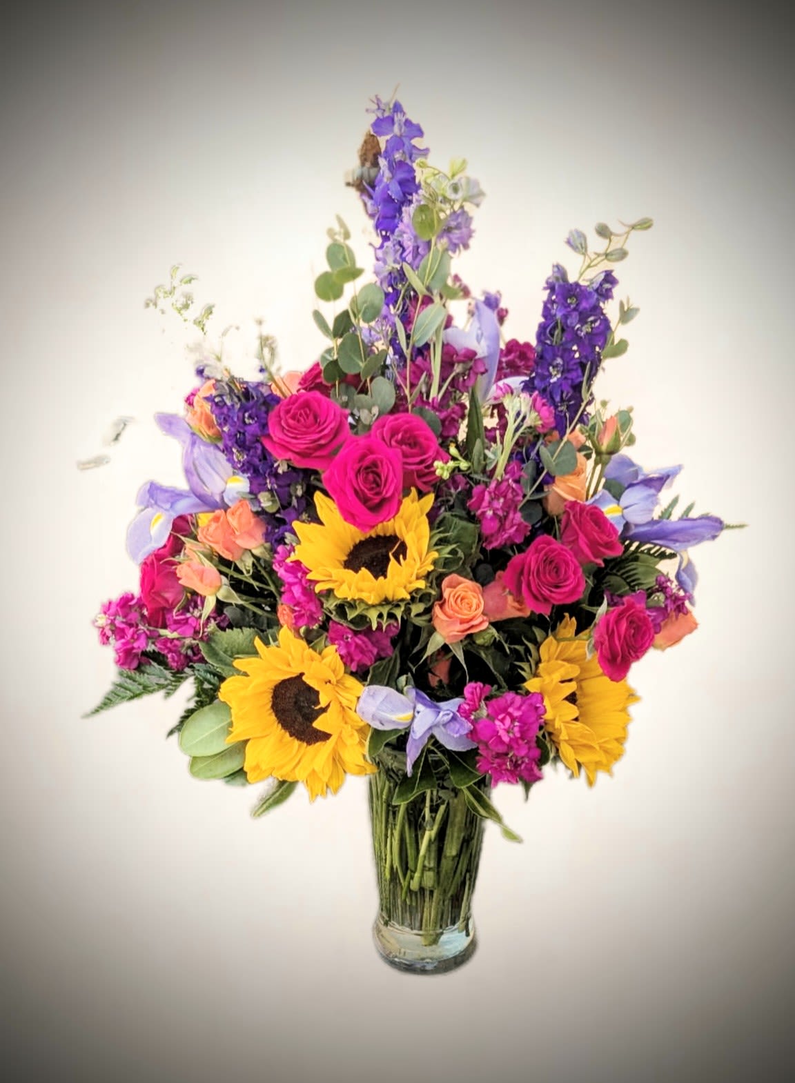 Summer Radiance Mixed Bouquet Summer Radiance Mixed Bouquet - Bursting with bold color and cheerful energy, our Summer Radiance arrangement is a celebration of sunshine and joy. This lively bouquet features brilliant sunflowers, fuchsia roses, delicate peach spray roses, and striking purple delphinium, artfully arranged with lush greenery and accents of soft lavender blooms. Designed in a clear glass vase, this tall and elegant arrangement is perfect for birthdays, celebrations, or simply brightening someone’s day. Perfect for: Summer gifts, special occasions, or adding a pop of color to any space.