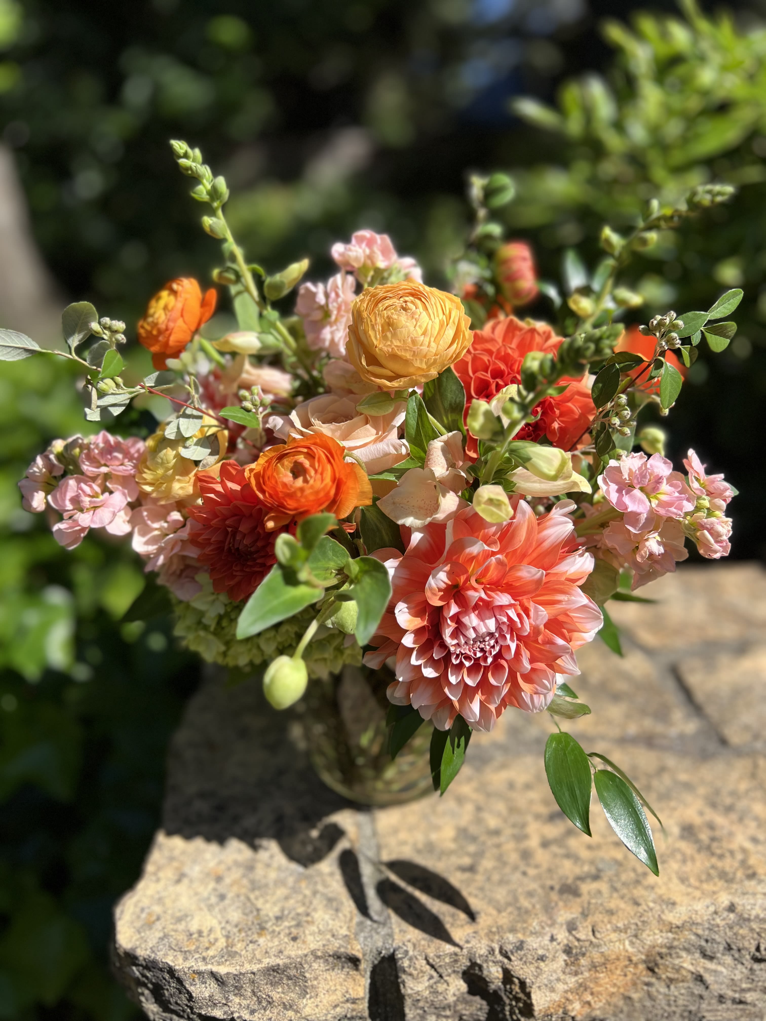 Orangina Orangina - All shades of orange for this summer cutie. Featuring dahlias, shimmer roses, ranunculus, and stock surrounded by green hydrangeas.