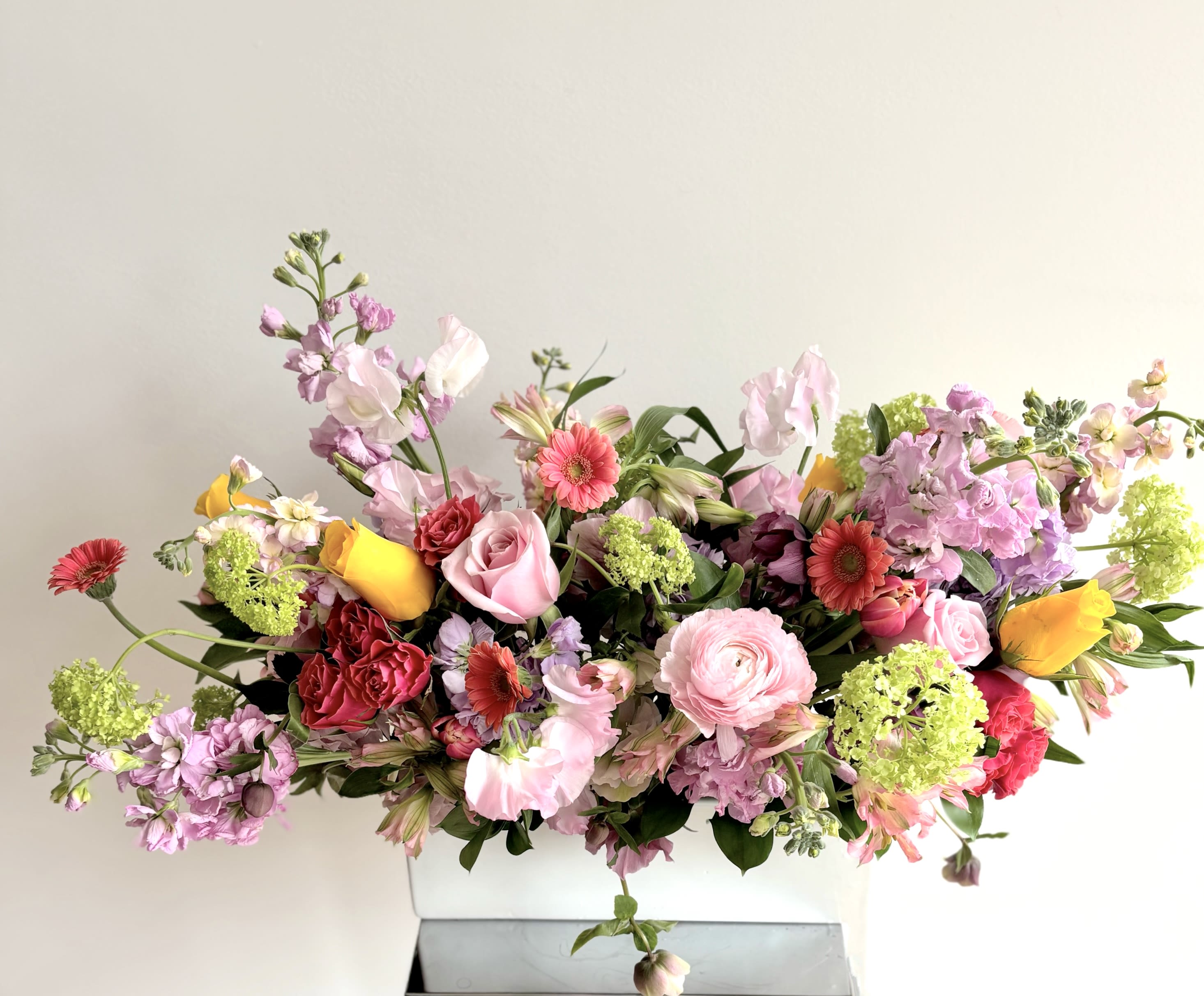 Spring In Bloom - An elegant long flower centerpiece in a white ceramic rectangular container. A lush melange of flowers with focus on spring flowers like viburnum, tulips, hyacinths, hellebore and sweet peas in addition to roses, stock, ranunculus, gerbers, lisiantnus and more. The color palette may vary based on the availability.