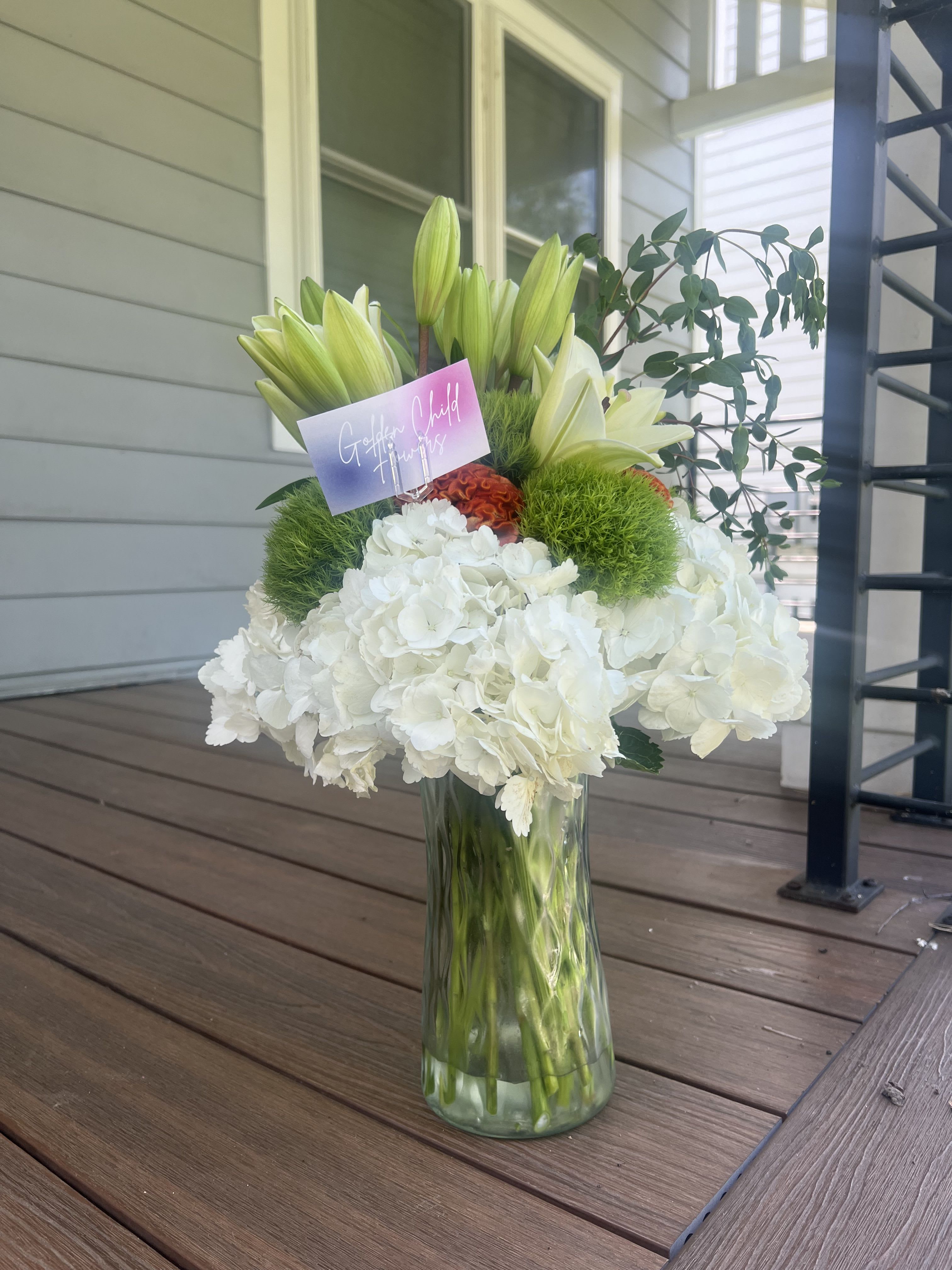 Color Me Happy Color Me Happy - A timeless mix of elegance and texture, this arrangement features lush white hydrangeas as the foundation, paired with crisp white oriental lilies just beginning to bloom. Vivid green trick dianthus (green poms) and a pop of orange cockscomb add playful contrast and dimension, while fresh eucalyptus stems lend movement and a natural finish. Designed in a clear glass vase, this piece is perfect for celebrations, sympathy, or simply bringing beauty into any space