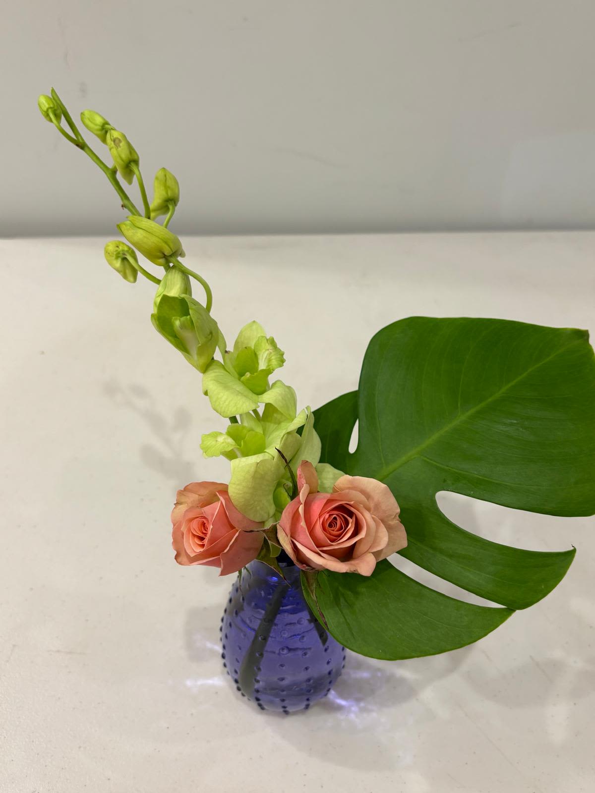 Tropical Minimalism Tropical Minimalism - A sleek and modern floral design combining soft peach roses, elegant green orchids, and a bold monstera leaf. Arranged in a textured violet vase, this minimalist composition brings a fresh, tropical touch with effortless sophistication — perfect for contemporary spaces