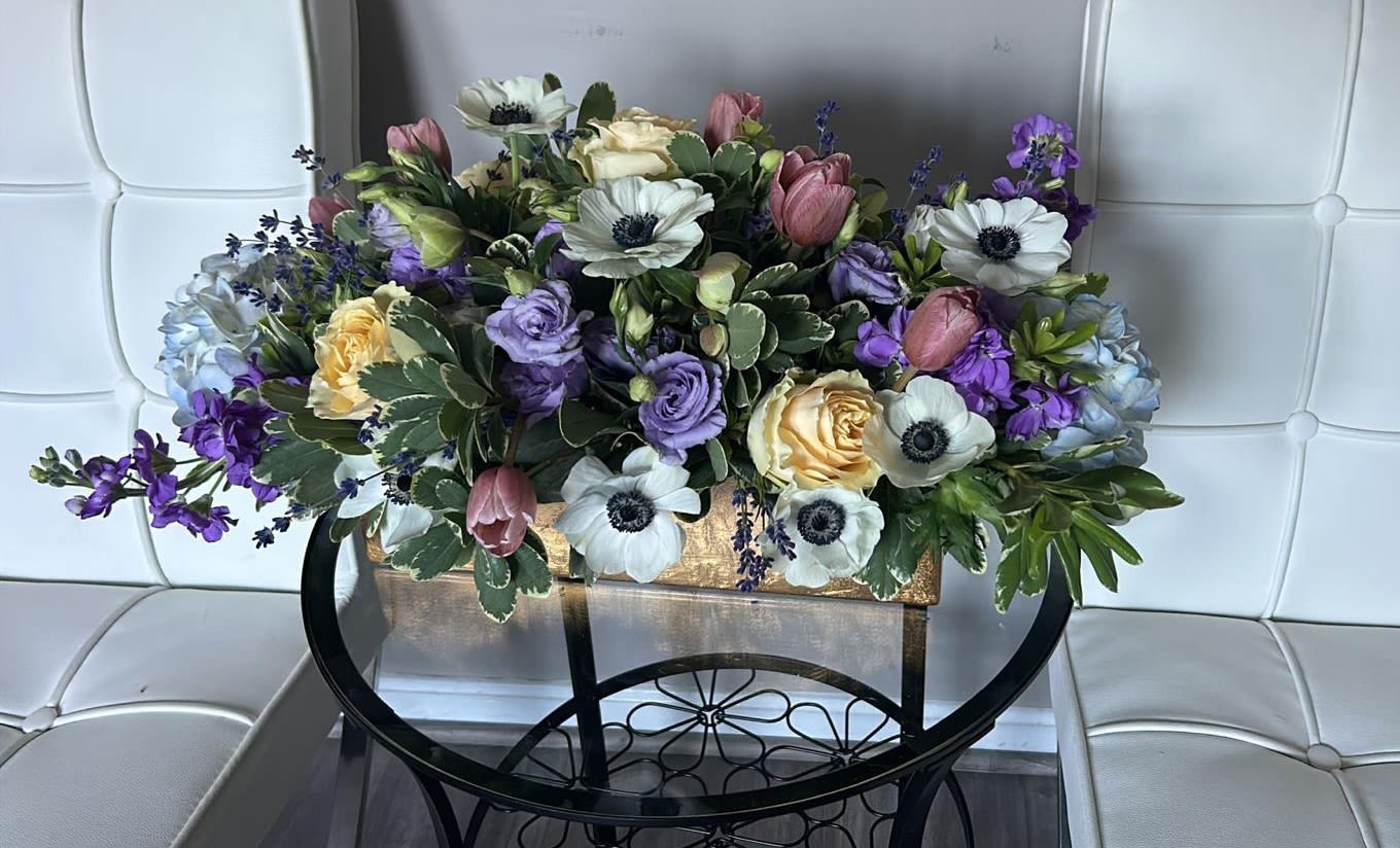 Vintage Elegance - A graceful and refined floral arrangement blending soft blush tulips, creamy garden roses, delicate anemones, lavender lisianthus, and accents of blue hydrangea. Framed by lush greenery and displayed in a textured gold container, this design evokes timeless sophistication — perfect for elegant events or classic home décor