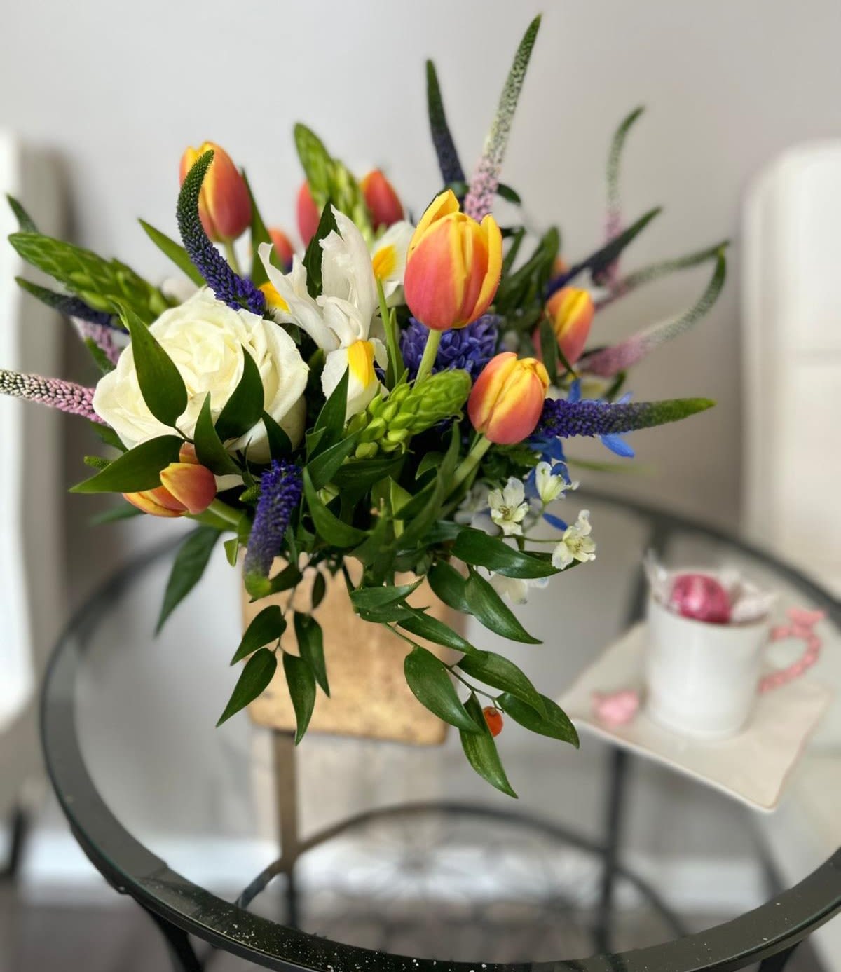 Spring Awakening Spring Awakening - A lively and vibrant arrangement bursting with energy, featuring two-tone tulips in fiery yellow and orange, pure white roses, delicate blue delphinium and playful sprigs of veronica. Accented with lush greenery and arranged in a modern gold vase, this cheerful design brings the joy and freshness of spring indoors
