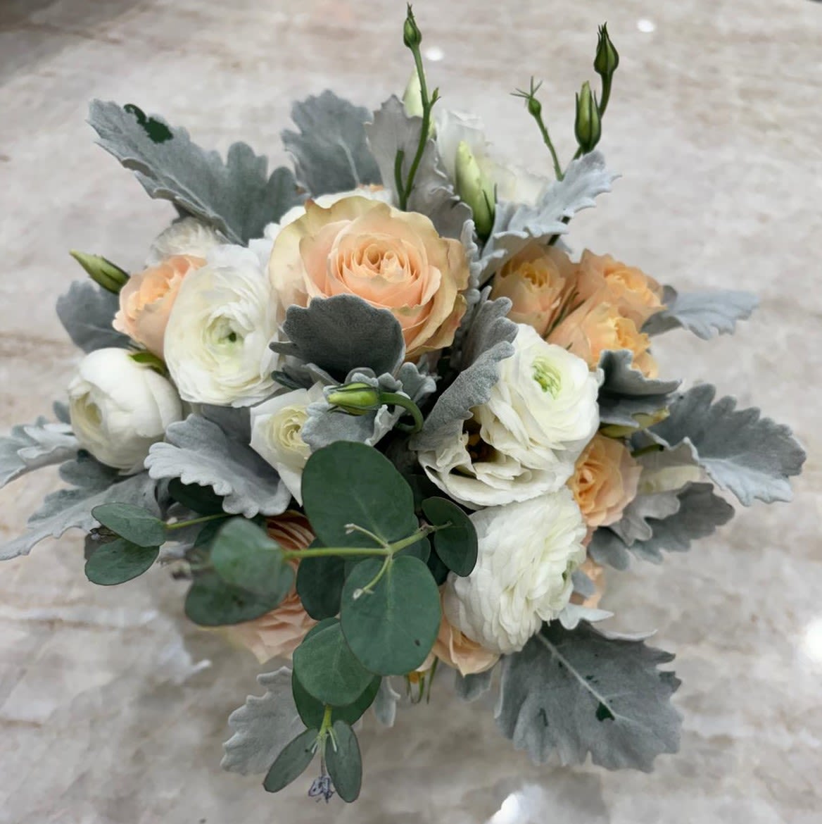 Soft Serenity - A delicate and elegant arrangement featuring soft peach roses, creamy ranunculus, and silvery dusty miller leaves, beautifully complemented by fresh eucalyptus. This design exudes calmness and understated sophistication — a perfect accent for intimate weddings, refined gatherings, or serene home décor