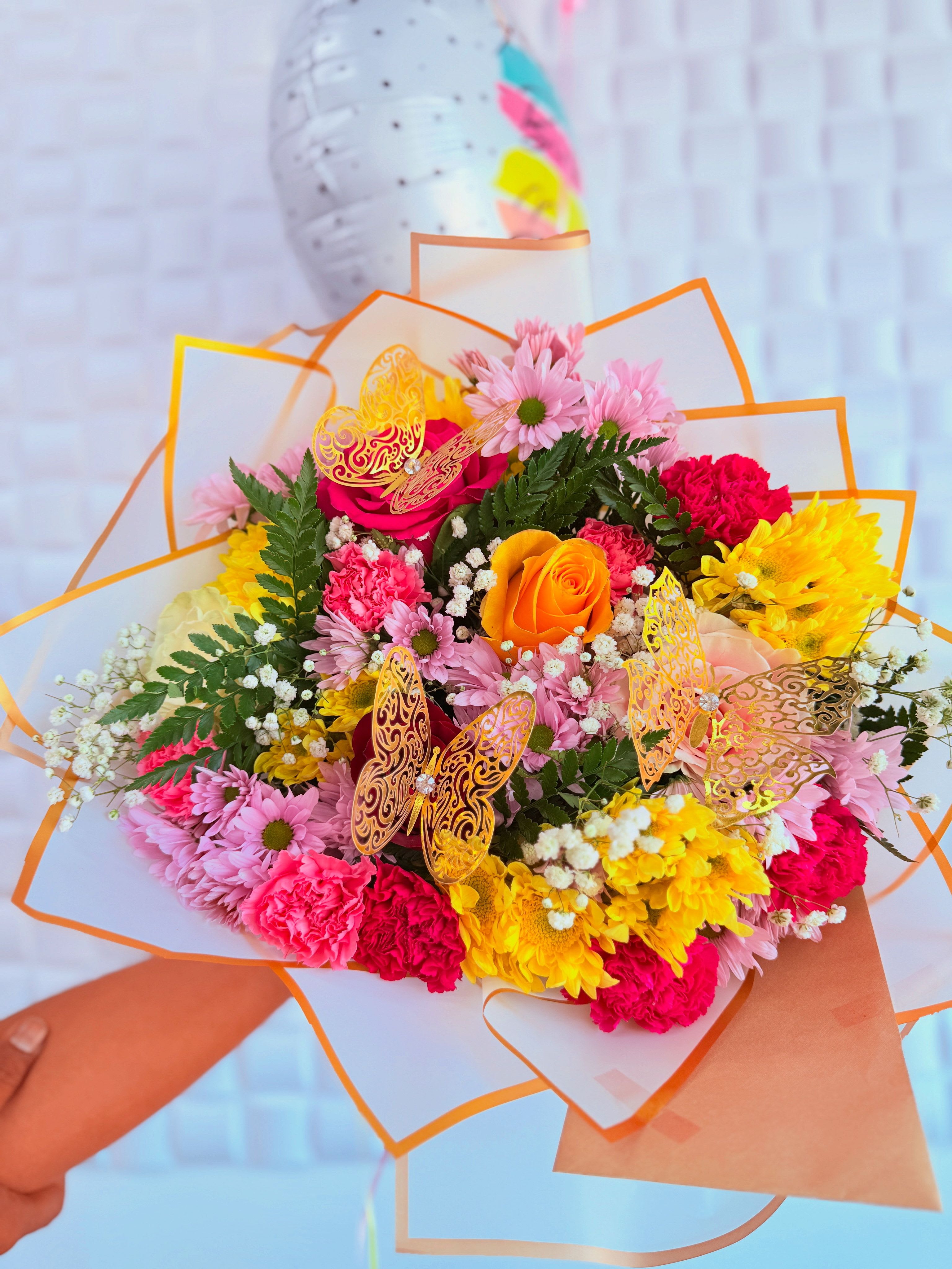 Enchanted Butterfly -  A radiant celebration of color and charm, this bouquet bursts with joyful energy and delicate artistry. At its heart lies an exquisite &quot;orange rose&quot;, symbolizing enthusiasm and admiration, gracefully surrounded by soft &quot;pink daisies&quot; and bold &quot;carnations&quot; in hues of crimson and blush. Cheerful &quot;yellow chrysanthemums&quot; add a sunny vibrance, evoking warmth and optimism.  Nestled among the florals are sprigs of &quot;baby’s breath&quot;, offering a gentle cloud-like contrast, while textured &quot;fern leaves&quot; lend lush greenery and movement. Two ornate &quot;golden butterflies&quot; delicately perched within the arrangement bring a whimsical and enchanting flourish—like nature's own jewelry adorning a masterpiece.  All of this is wrapped in crisp &quot;white paper with orange accents&quot;, elegantly framing the ensemble and tying together the palette in a modern yet timeless presentation. A playful &quot;balloon in the background&quot; suggests celebration—perhaps a birthday or a heartfelt occasion—making this bouquet a perfect expression of joy, appreciation, or simply love in bloom. 