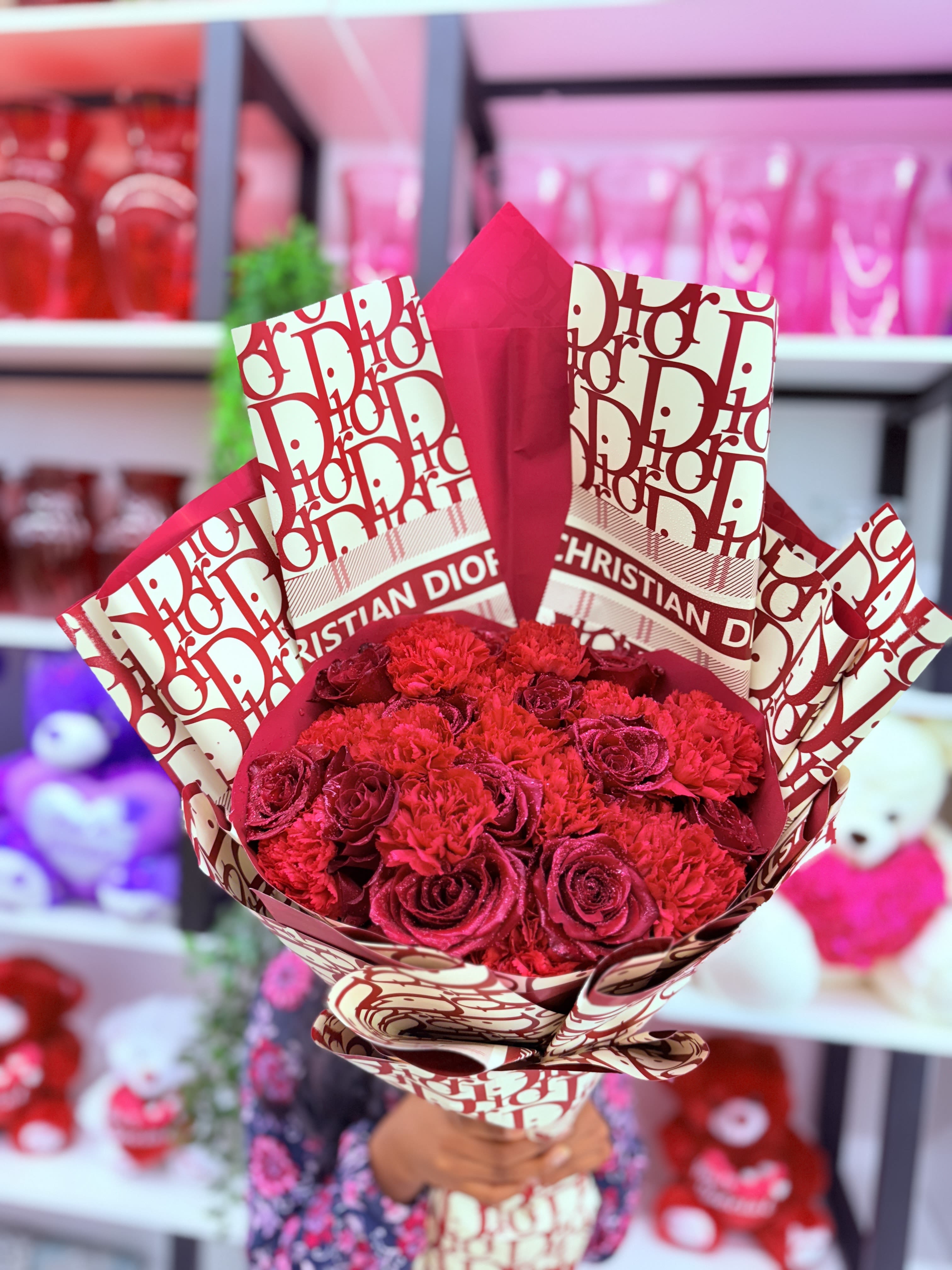 Blooming Red -  A true emblem of passion and high fashion, this bouquet stuns with its rich **monochromatic palette of deep red roses and carnations**, artfully arranged and elegantly wrapped in **signature Christian Dior–branded paper**. The red blooms exude classic romance and timeless beauty—**roses** symbolizing deep love and desire, while **carnations** add texture and a graceful ruffle that enhances their allure.  The soft layering of petals creates a lush, full-bodied arrangement that feels both bold and intimate. Set against the sleek, branded wrapping with **golden Dior lettering**, the bouquet transcends traditional floral design—**it feels like a couture statement**, designed to impress and captivate.  Whether given in celebration or as a passionate gesture, this bouquet communicates sophistication, confidence, and enduring affection. It’s not just a floral gift—it’s a fashion-forward expression of emotion. 
