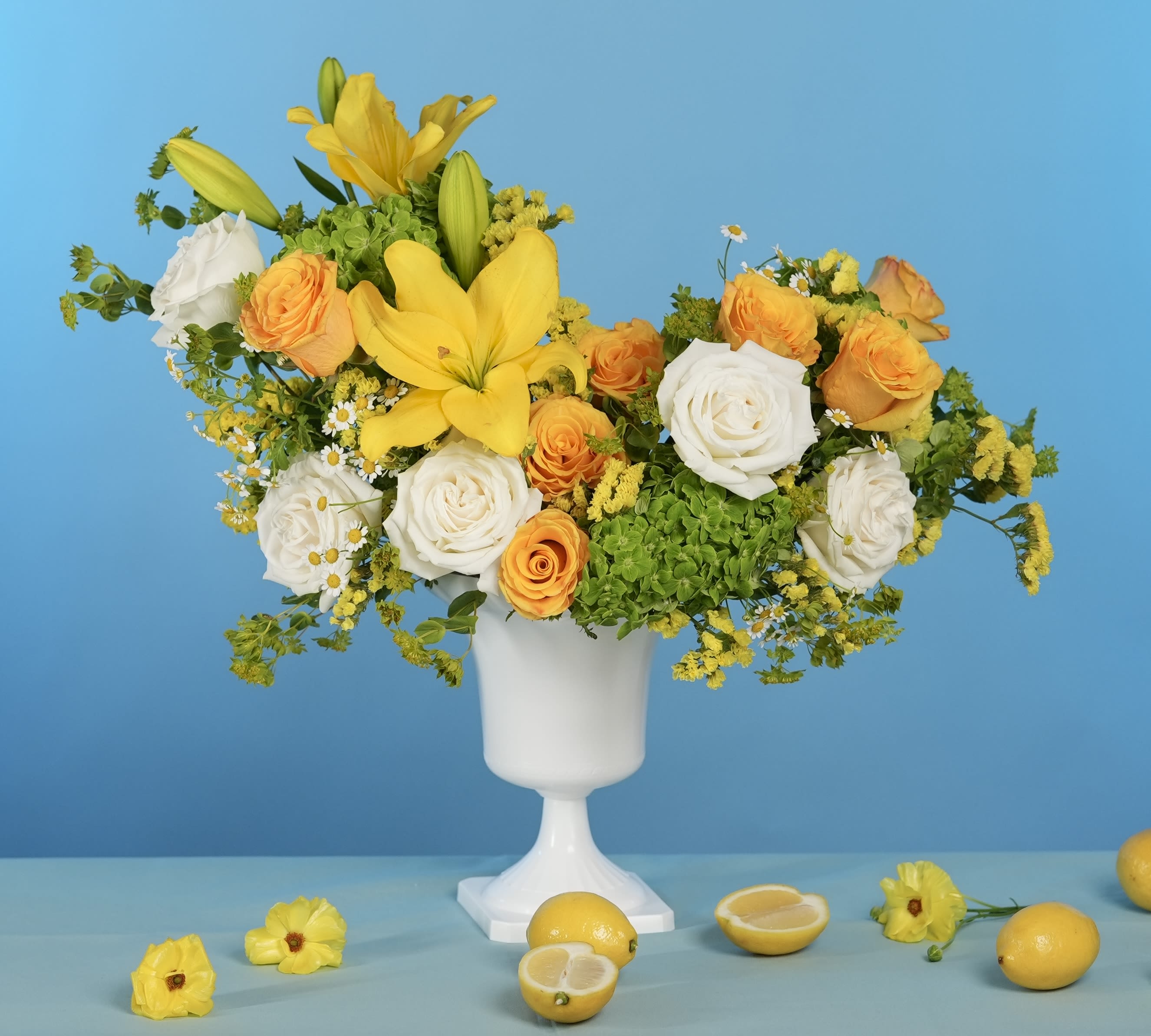 Summer Muse - Bask in the warmth of seasonal beauty with Summer Muse, a radiant arrangement that captures the golden essence of long, sunlit days. Designed in a crisp white pedestal vase, this piece blends buttery yellow lilies, white garden roses, cheerful orange roses, soft chamomile, and green hydrangeas with natural movement and charm.  Perfect for summer celebrations, elegant gatherings, or simply brightening your space with joyful sophistication.  Style: Bright, elegant, uplifting Fragrance: Light citrus and soft floral notes