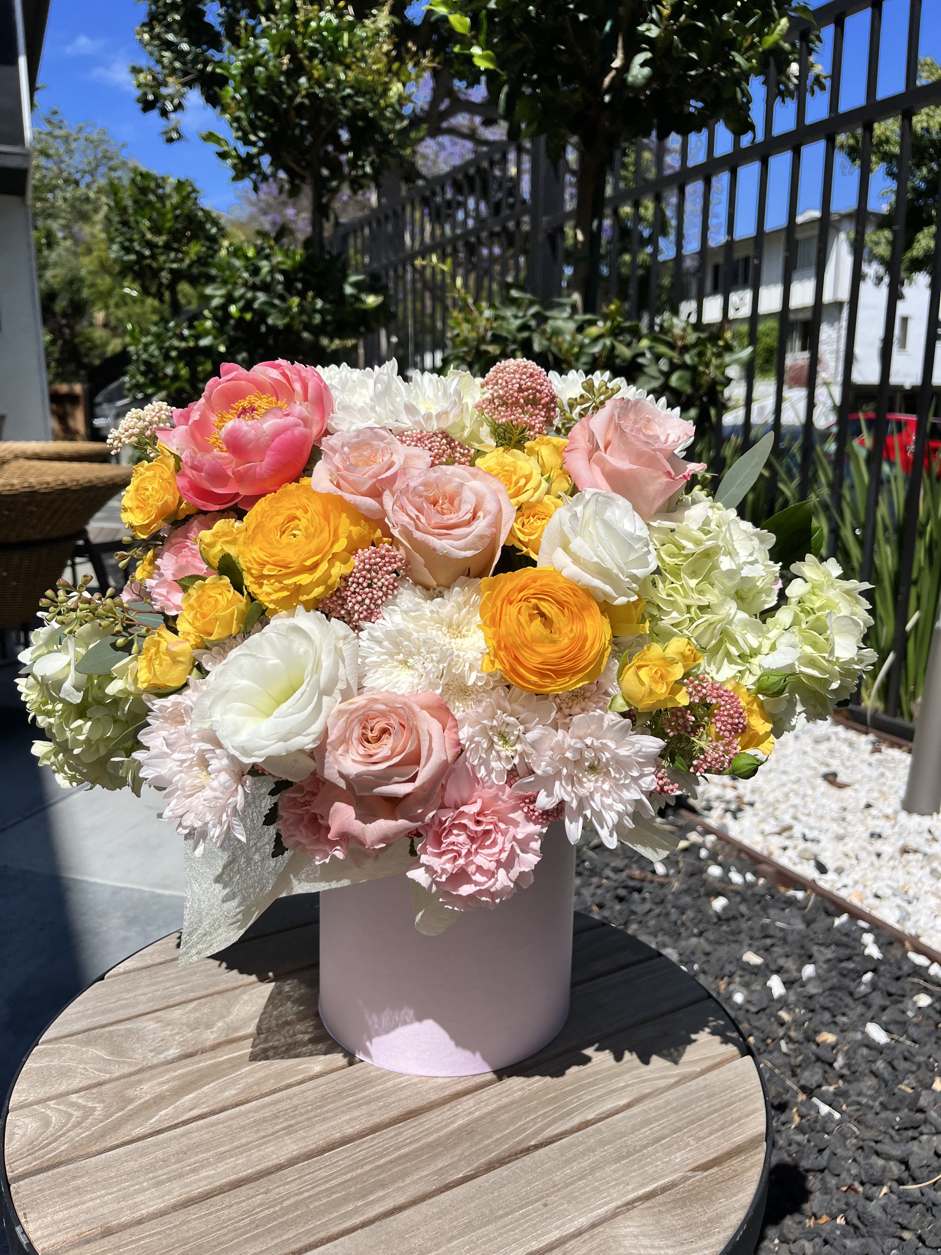Golden Blush - Golden Blush – Designer Hatbox Arrangement Radiant and graceful, Golden Blush is a luminous floral creation that captures the glow of golden hour in full bloom. Nestled in a soft pink hatbox, this bouquet blends sunshine-yellow ranunculus, soft blush roses, creamy lisianthus, vibrant peonies, and airy chrysanthemums with touches of hydrangea and yarrow. The result is a harmonious burst of color that feels fresh, refined, and full of life.  Perfect for birthdays, spring celebrations, or moments that call for joy and elegance.  Style: Elegant, cheerful, radiant Fragrance: Lightly floral with fresh garden notes 