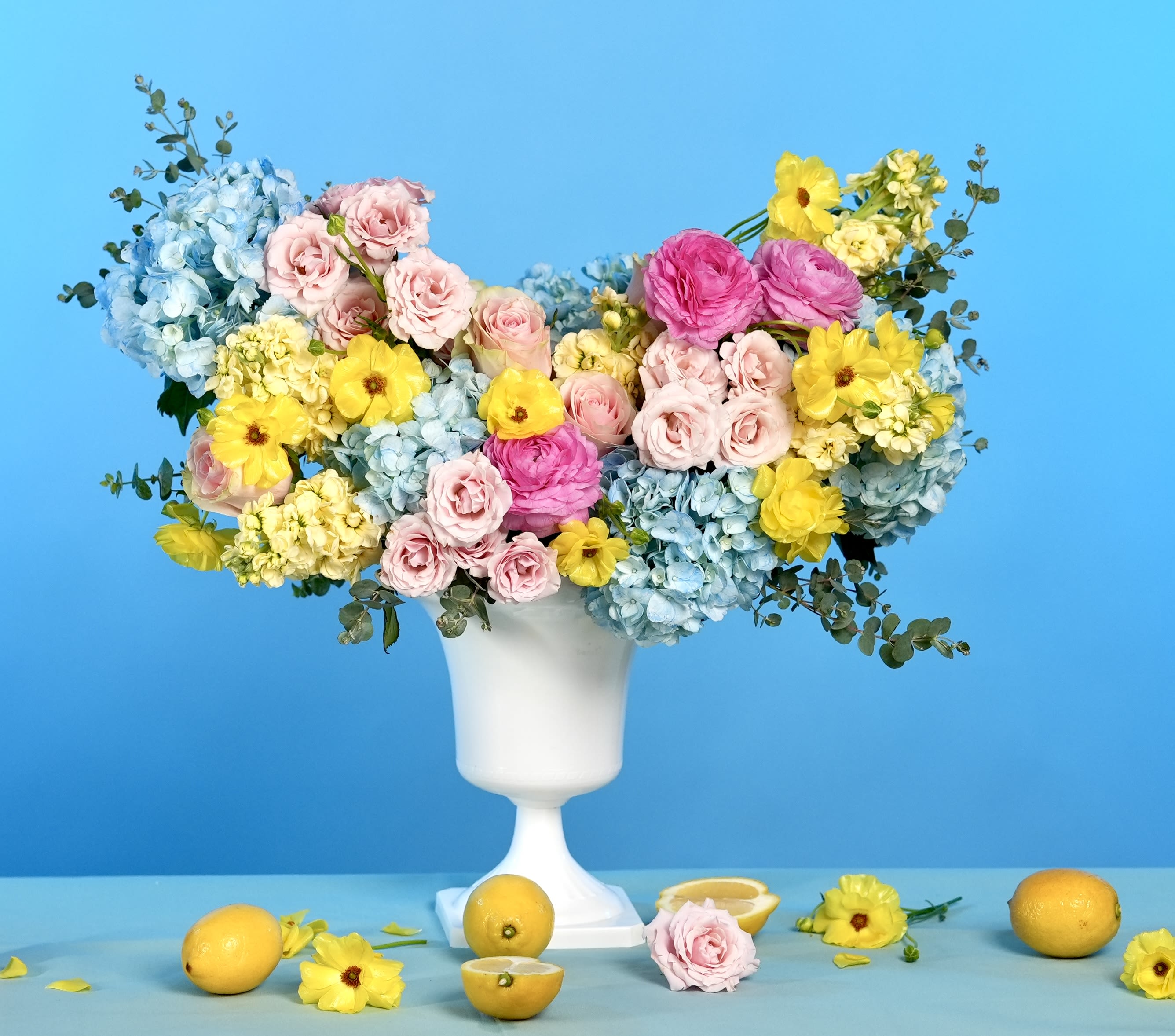Elizabeth's Elegance - Regal, radiant, and gracefully composed, Elizabeth’s Elegance is a floral ode to timeless beauty. This artfully designed arrangement features airy blue hydrangeas, delicate blush roses, sunny yellow daffodils and ranunculus, pale lisianthus, and striking pink ranunculus — all styled in a tall white pedestal vase for a truly elevated presentation.  Reminiscent of royal gardens kissed by morning light, this bouquet is perfect for elegant events, refined celebrations, or a grand gesture of admiration.  Style: Classic, opulent, soft-glam Fragrance: Light garden floral with soft citrus notes