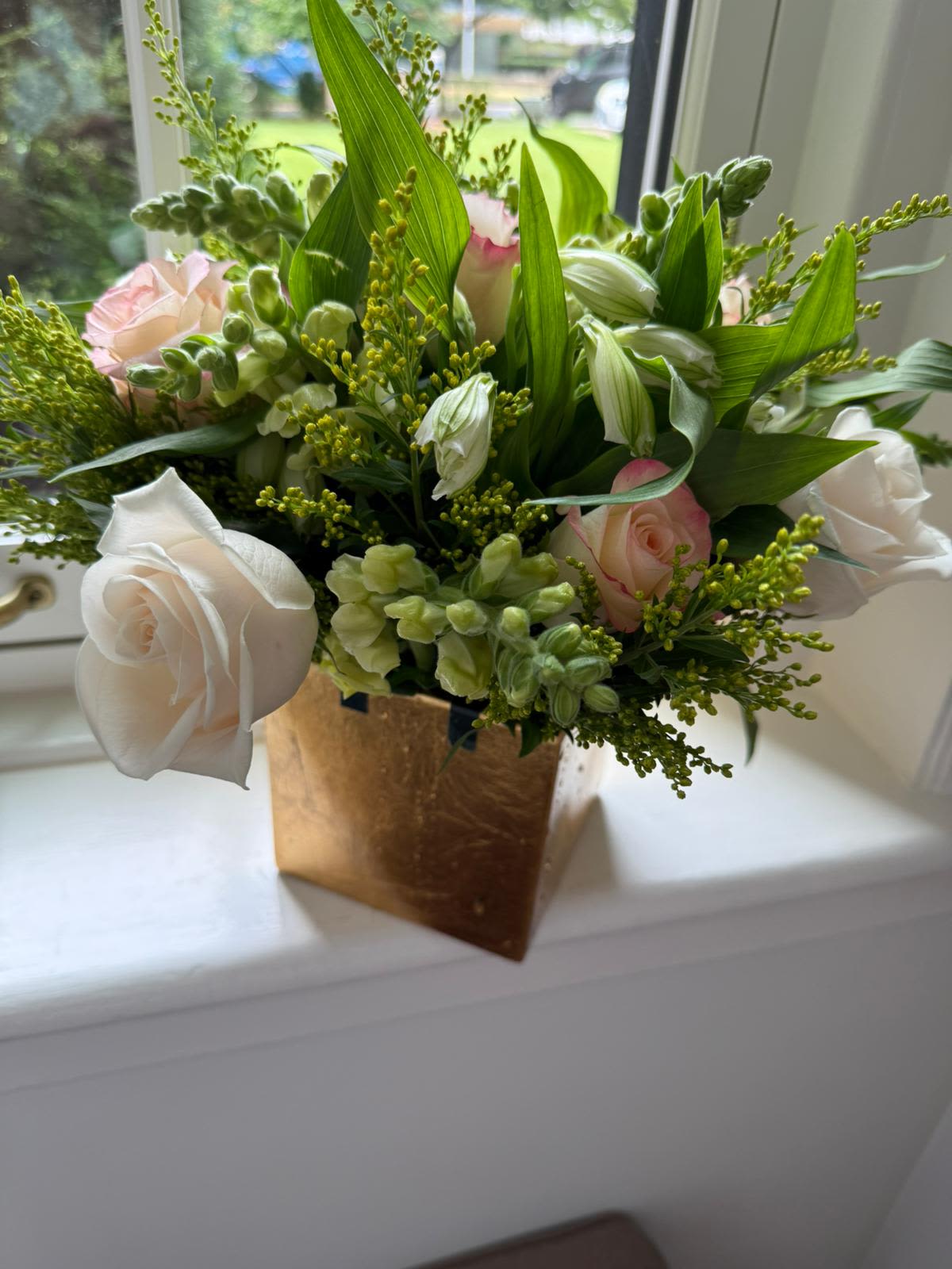 Whispering Petals - A soft and elegant arrangement of blush pink and ivory roses, creamy snapdragons, and white alstroemeria, delicately embraced by vibrant greenery and golden solidago. Set in a chic gold cube vase, this design radiates quiet grace — ideal for serene spaces, heartfelt gifts, or gentle morning light