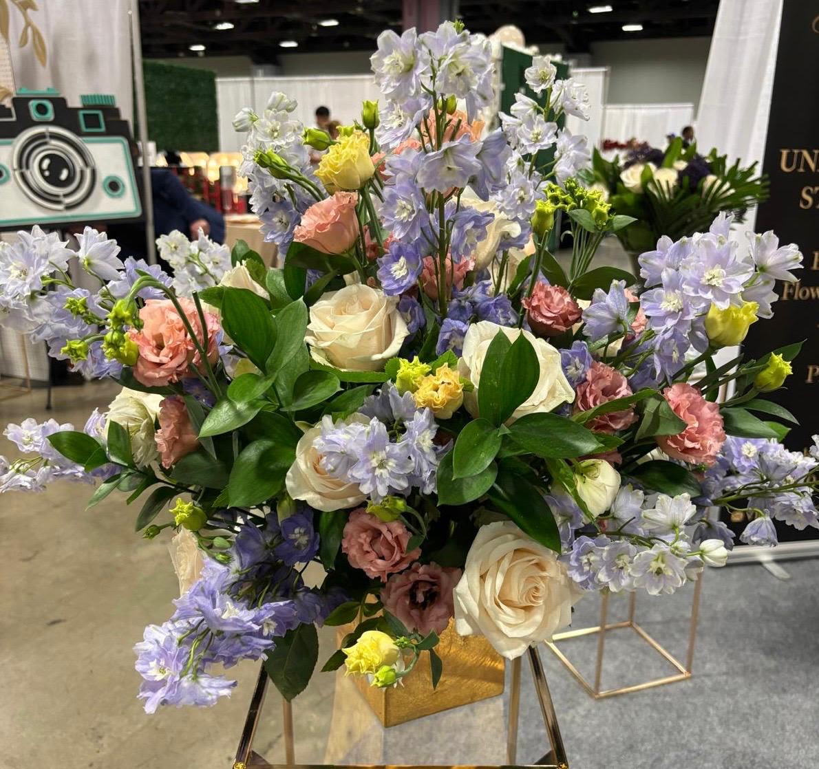 Pastel Symphony Pastel Symphony - A graceful and airy arrangement bursting with soft pastel hues — featuring creamy ivory roses, pale lavender delphiniums, blush lisianthus, and sunny hints of yellow. Accented with fresh greenery and displayed in a chic gold vase, this bouquet exudes romantic charm and gentle elegance, perfect for spring celebrations or timeless decor