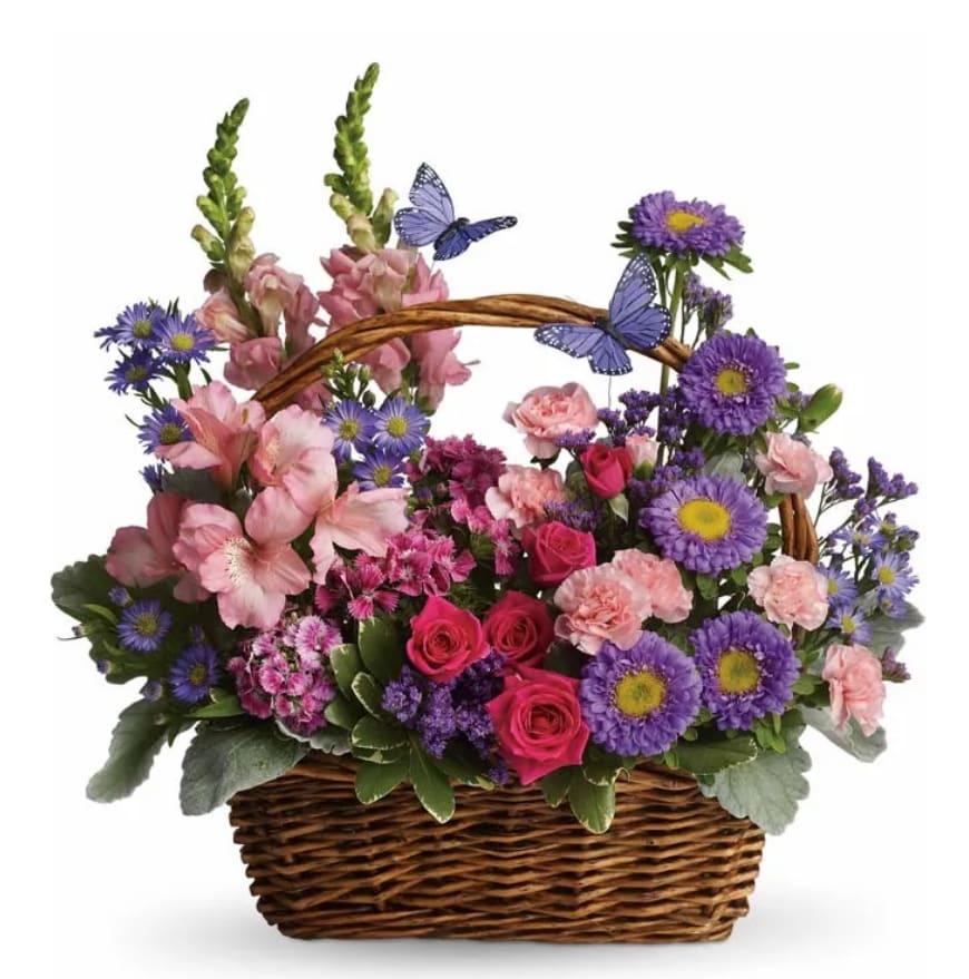 Country Basket Blooms - Step into the serenity of a blooming country meadow with this charming floral basket. A delightful mix of soft pink and rich purple blossoms nestles gracefully within a woven natural basket, capturing the spirit of a sunny countryside stroll. Above, whimsical purple butterflies seem to dance in the air, adding a playful and magical touch. Whether it's to brighten a room or lift someone's spirits, this lovely arrangement brings a breath of fresh meadow air to any heart or home.