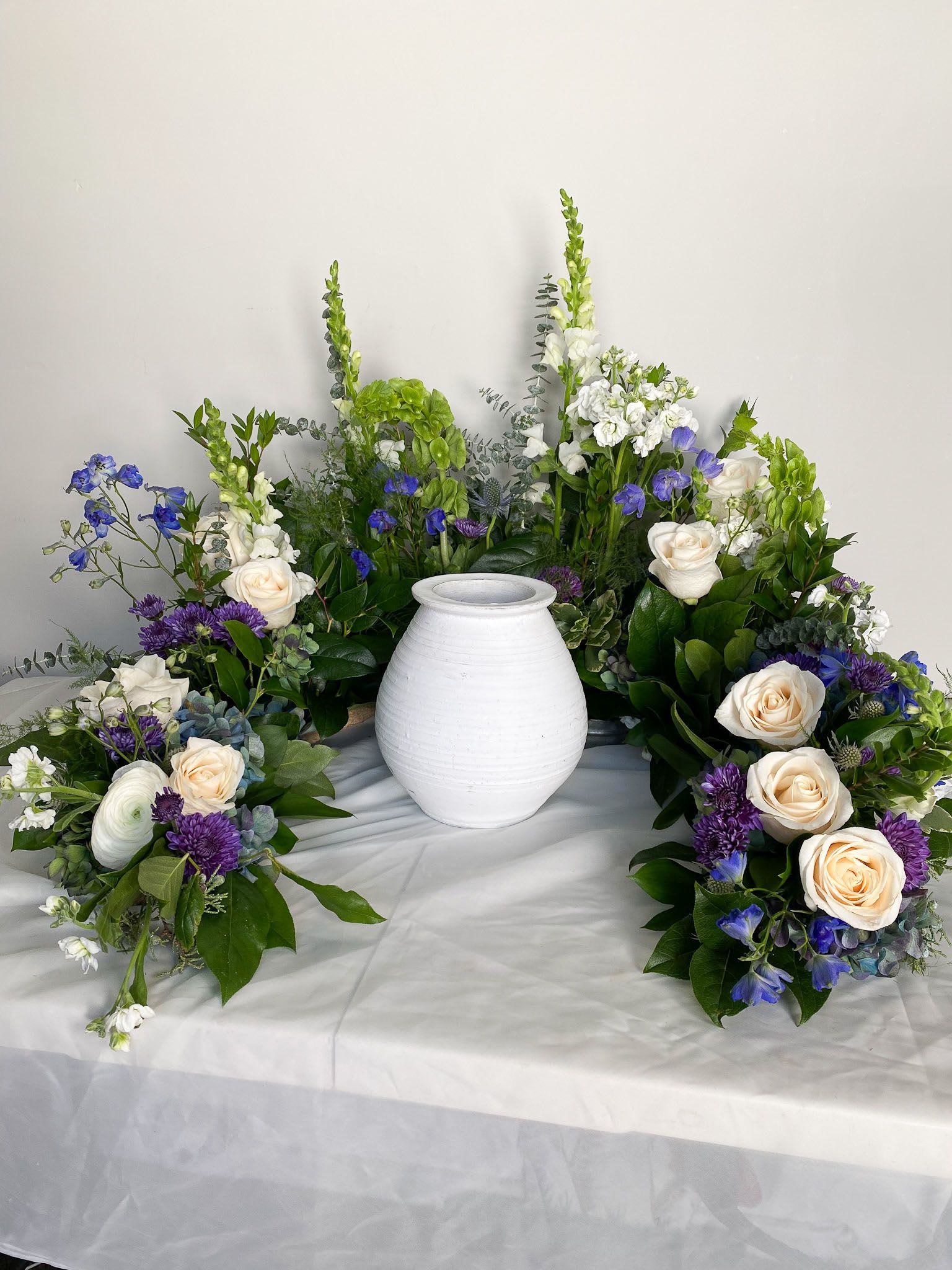 Encircling Memorial - Honor cherished memories with a graceful floral arrangement designed to encircle an urn, symbolizing eternal love and remembrance. This thoughtful piece features a harmonious blend of lush greenery and seasonal blooms, carefully chosen for their timeless beauty and symbolic meaning.
