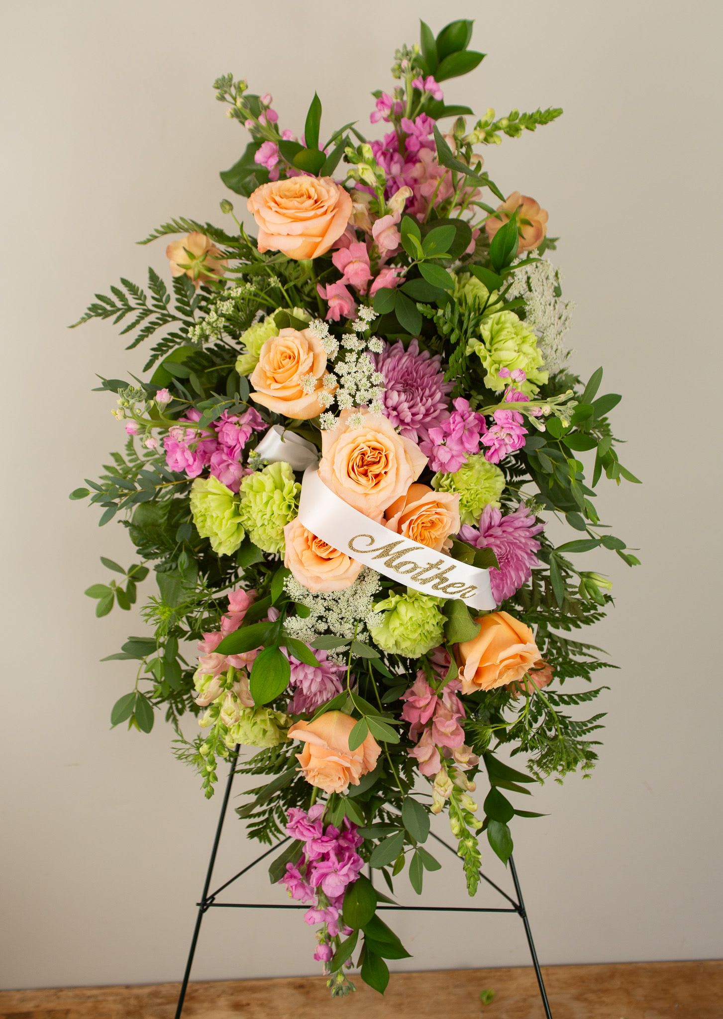 Pink Traditions Spray - A graceful tribute in soft, comforting hues. This standing spray features a blend of pink and warm-toned blooms, thoughtfully accented with fresh greenery. Designed to honor and celebrate a loved one’s life, each arrangement is made with seasonal flowers—selections may vary to reflect your preferences and what’s most beautiful at the time.  This is customizable to accommodate more pink hues as desired. 