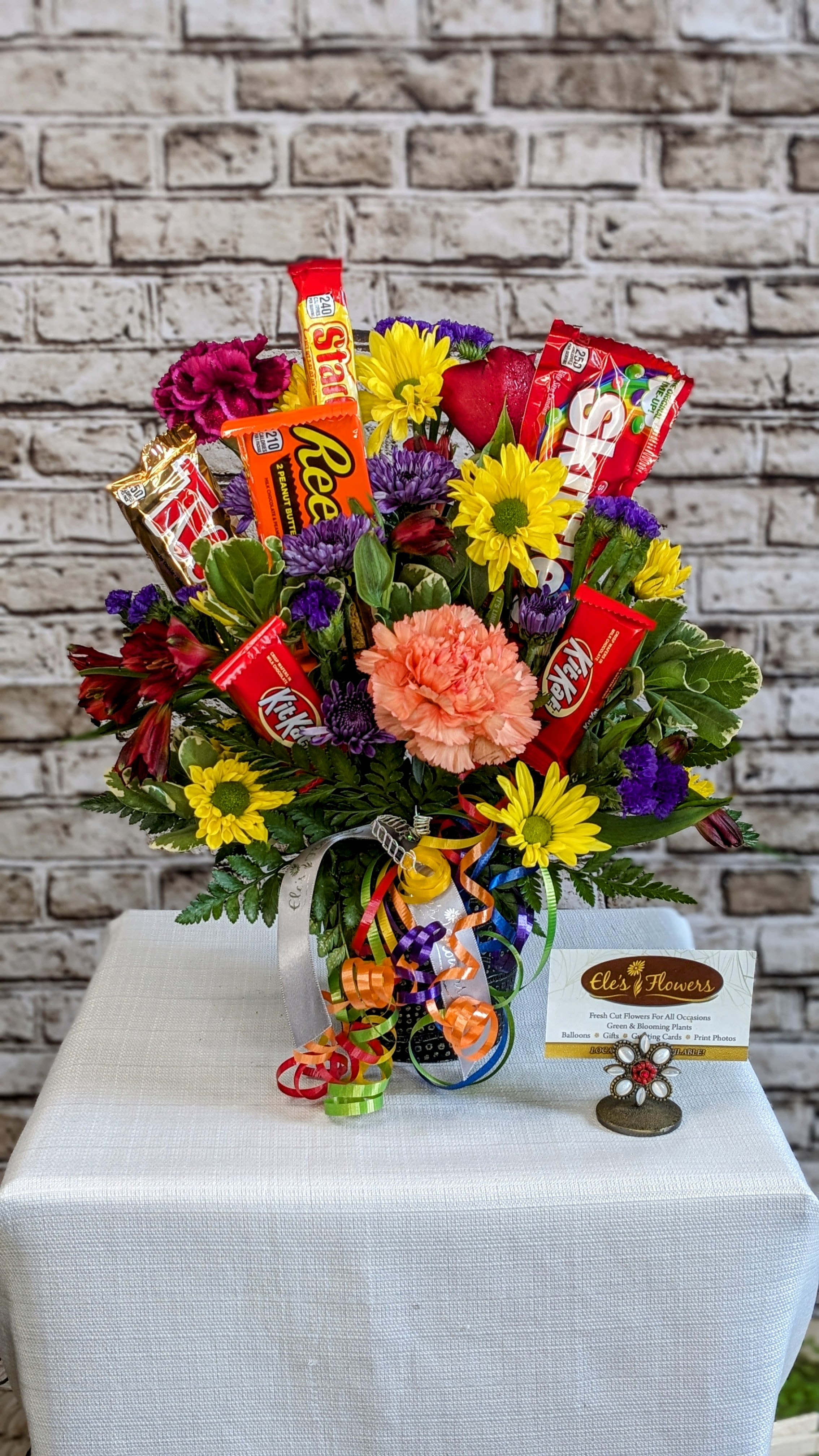 Confectionary Garden - This vibrant and joyful arrangement features a stunning mix of bright, rich, and bold primary color flowers that pop with energy and warmth. Adorned with an assortment of sweet candy bars and treats, it offers a delightful treat for both the eyes and the taste buds. Nestled in a container or vessel, the arrangement is complemented by fresh, lush foliage, adding a natural, verdant touch. Perfect for celebrations such as birthdays, congratulations, get well wishes, or simply as a cheerful &quot;just because&quot; gesture. Please note that blooms and sweet treats may vary based on availability, ensuring a unique and fresh touch in every arrangement.  Approximate Dimensions: 16&quot;H x 13&quot;W