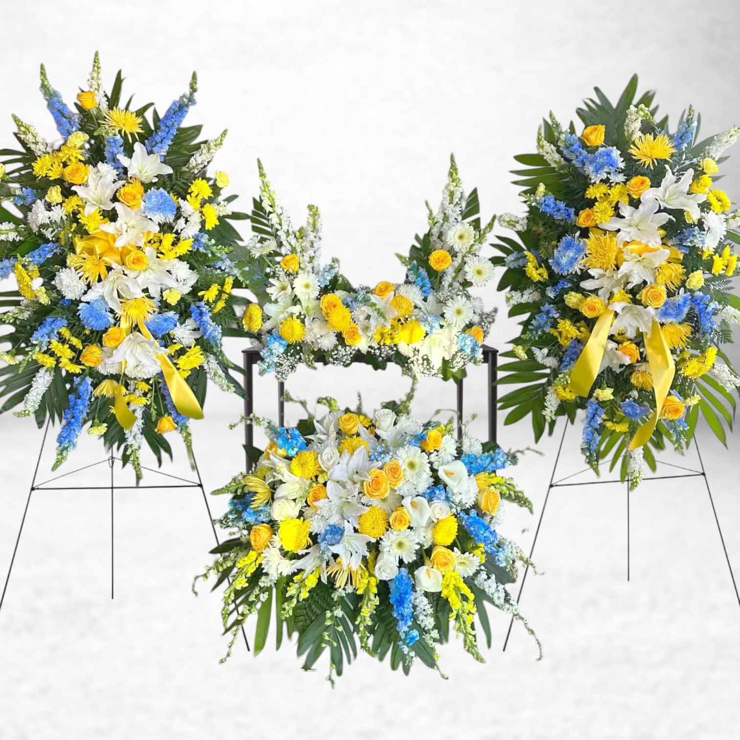 Sunrise Remembrance Collection - The Sunrise Remembrance Collection is a radiant expression of peace, hope, and honor. This coordinated five-piece sympathy set includes two standing sprays, two pedestal arrangements, and one lush casket spray—each featuring a vibrant blend of yellow roses, blue delphinium, white lilies, chrysanthemums, and snapdragons accented with tropical greenery and satin ribbon.  This collection is ideal for honoring a life well-lived, offering warmth and solace at the funeral home, church, or graveside service. Whether you're remembering a loved one with a sunny disposition or celebrating a life full of light and joy, these arrangements provide a comforting tribute to a beautiful legacy.  Lovingly crafted by Twigs and Blooms in Galveston, we offer same-day funeral flower delivery to all local Galveston funeral homes, including those in Friendswood, League City, Dickinson, Texas City, and the greater Texas Gulf Coast area.