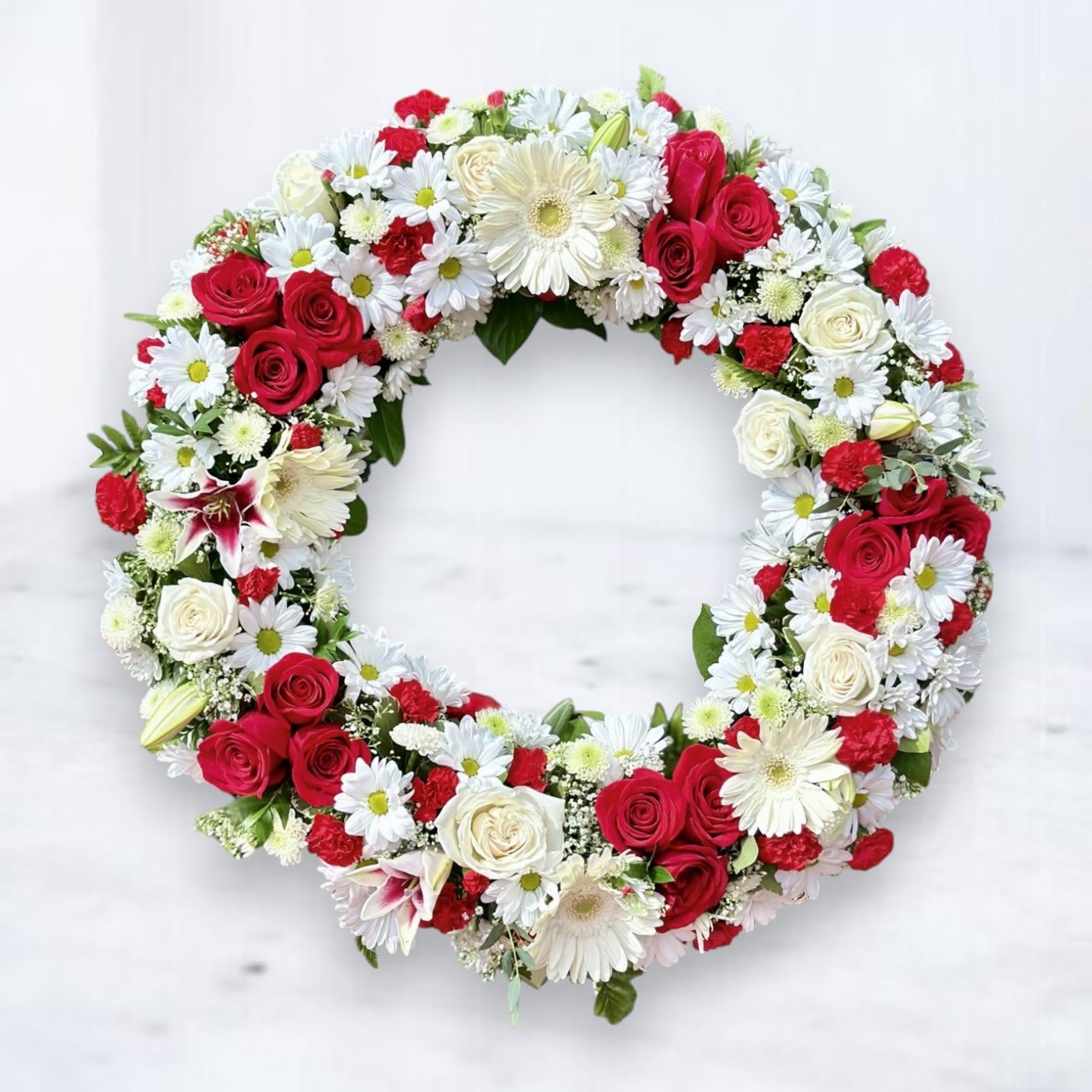 Blossoms of Honor Wreath - Pay tribute to a cherished life with the &quot;Blossoms of Honor Wreath,&quot; a stunning and heartfelt arrangement crafted to convey deep respect and remembrance. This elegant funeral wreath features a harmonious blend of red roses symbolizing love and devotion, white roses and daisies representing purity and grace, and delicate carnations for admiration. Each bloom is thoughtfully arranged in a classic circular shape, signifying the continuity of life and the eternal bond shared with loved ones.  Easels are included in the price of all wreath arrangements to ensure easy display at services.