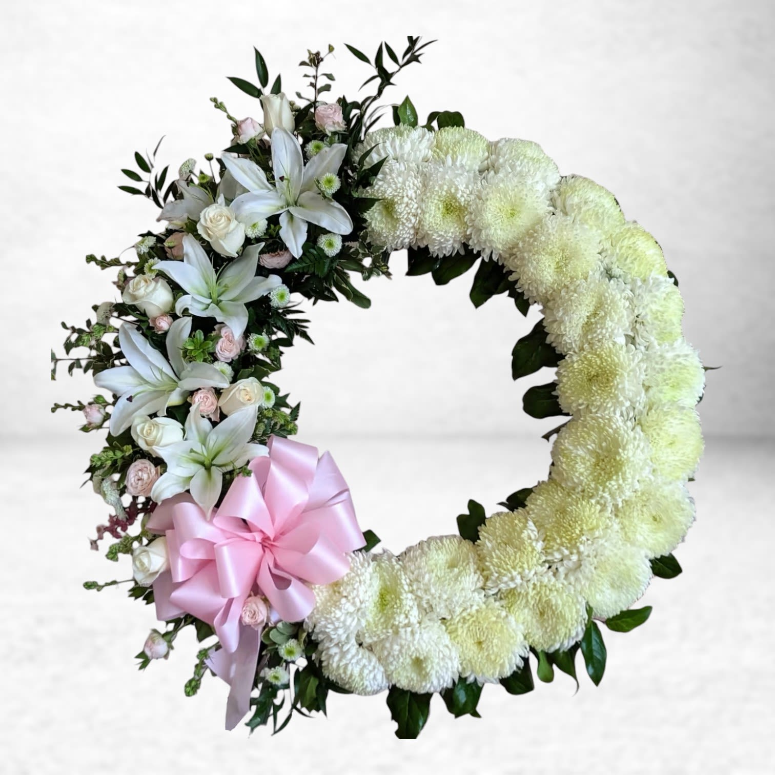 Heaven’s Embrace Funeral Wreath - The Heaven’s Embrace Wreath is a graceful expression of peace and remembrance, featuring pristine white chrysanthemums, soft blush spray roses, and pure white lilies arranged in a refined crescent design. A satin pink bow completes the tribute with a touch of gentle color and elegance.  This wreath is a beautiful symbol of eternal love and serenity, ideal for honoring a mother, daughter, grandmother, or cherished friend at a memorial, funeral, or cremation service.  Lovingly crafted by Twigs and Blooms, your premier Galveston florist, we offer same-day sympathy flower delivery to local funeral homes, cemeteries, and residences throughout Galveston, Friendswood, League City, Dickinson, and the surrounding Texas Gulf Coast communities.