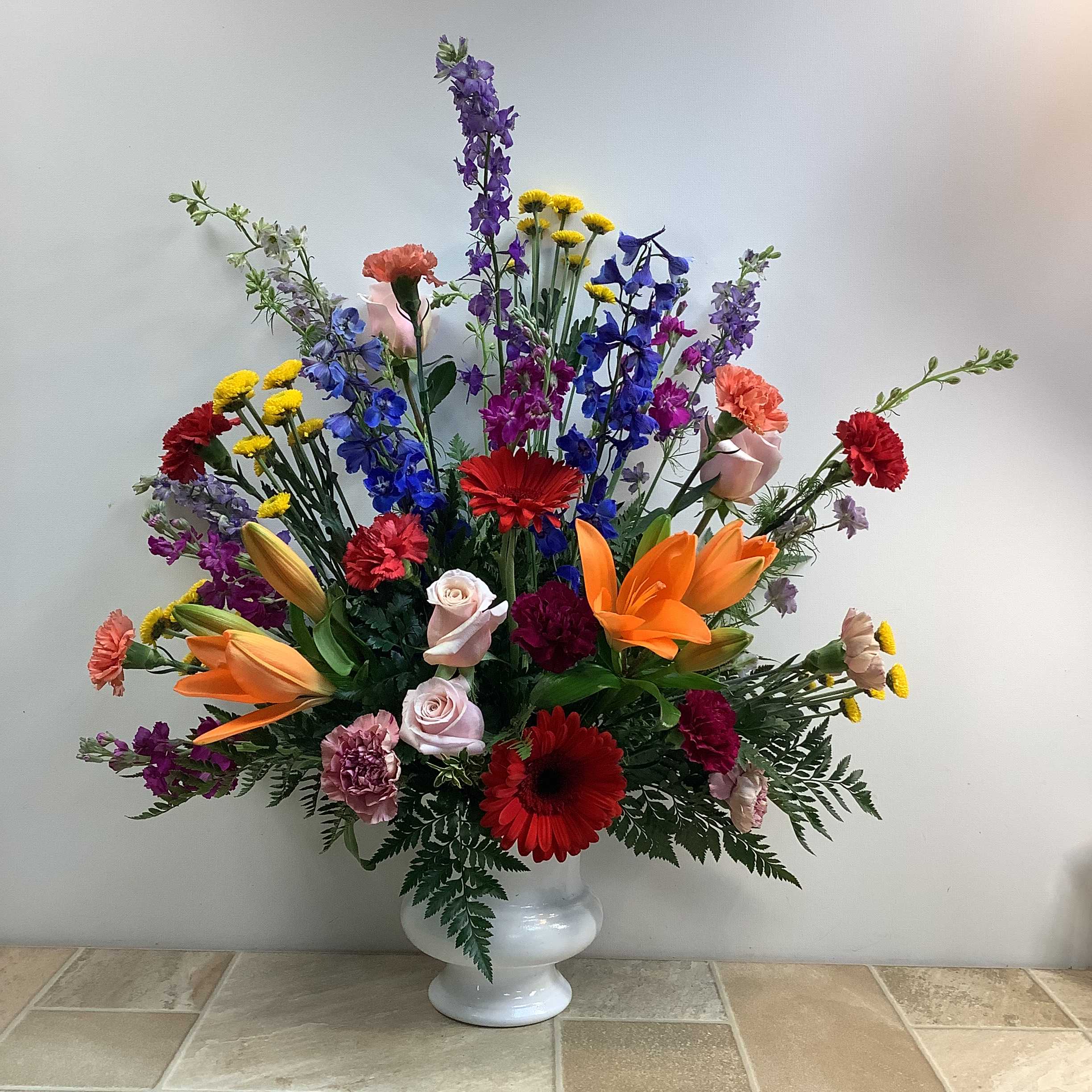Samuel Samuel - This stunning bouquet is a vibrant, large-scale design that features a diverse and colorful mix of flowers, artistically displayed in a white pedestal vase. Flowers include: bold and bright orange asiatic lilies, cheerful red gerbera daisies, elegant pink roses, textural red and pink carnations, whimsical yellow button mums, and flowy lavender larkspur and blue delphinium. Purple stock and feathery ferns and other greenery adds to the lushness of this bold arrangement. Primary elements: lily, daisy, rose, carnation, mum, fern