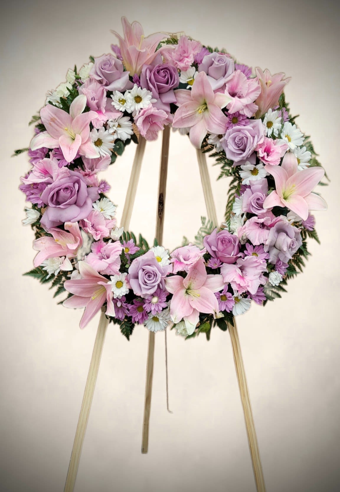 Peaceful Remembrance Wreath  - Soft, elegant, and heartfelt, the Peaceful Remembrance standing wreath offers a gentle tribute to a cherished life. Featuring blush pink lilies, lavender roses, white daisies, and delicate accents of pastel carnations and mums, this wreath radiates serenity and love. Artfully arranged with lush greenery on a wooden easel, it brings comfort and beauty to memorial services, funerals, or celebrations of life.  A timeless expression of sympathy and grace.   
