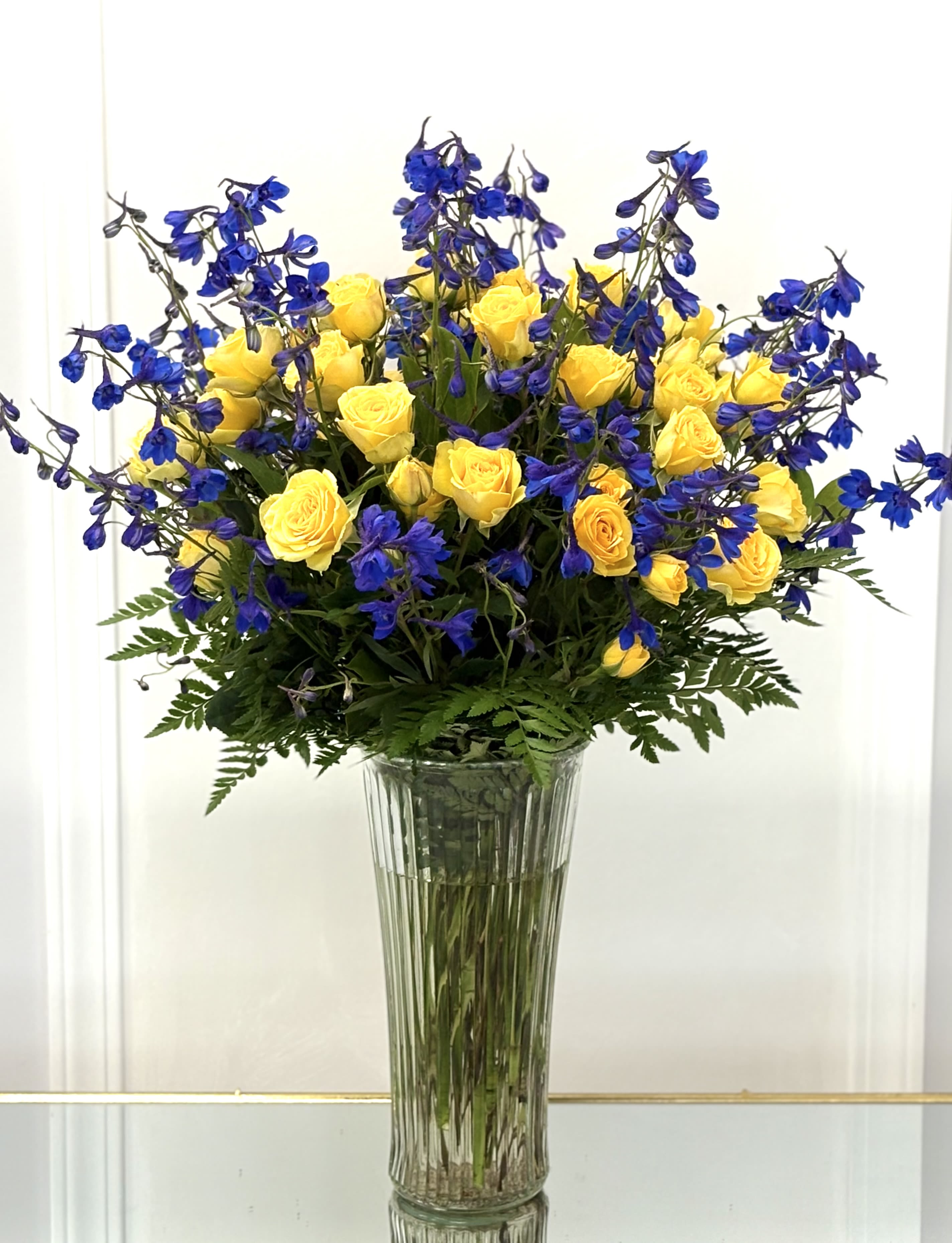 Golden Horizon By VLM Flowers - A radiant combination of sunshine-yellow spray roses and striking blue delphinium blooms, this tall glass vase arrangement is bursting with energy and contrast. The cheerful yellow symbolizes joy and friendship, while the deep blue adds a sense of depth and elegance. Lush greenery completes the composition, giving it a full and flourishing look. Perfect for celebratory occasions, grand gestures, or uplifting someone's day with the colors of optimism and strength.