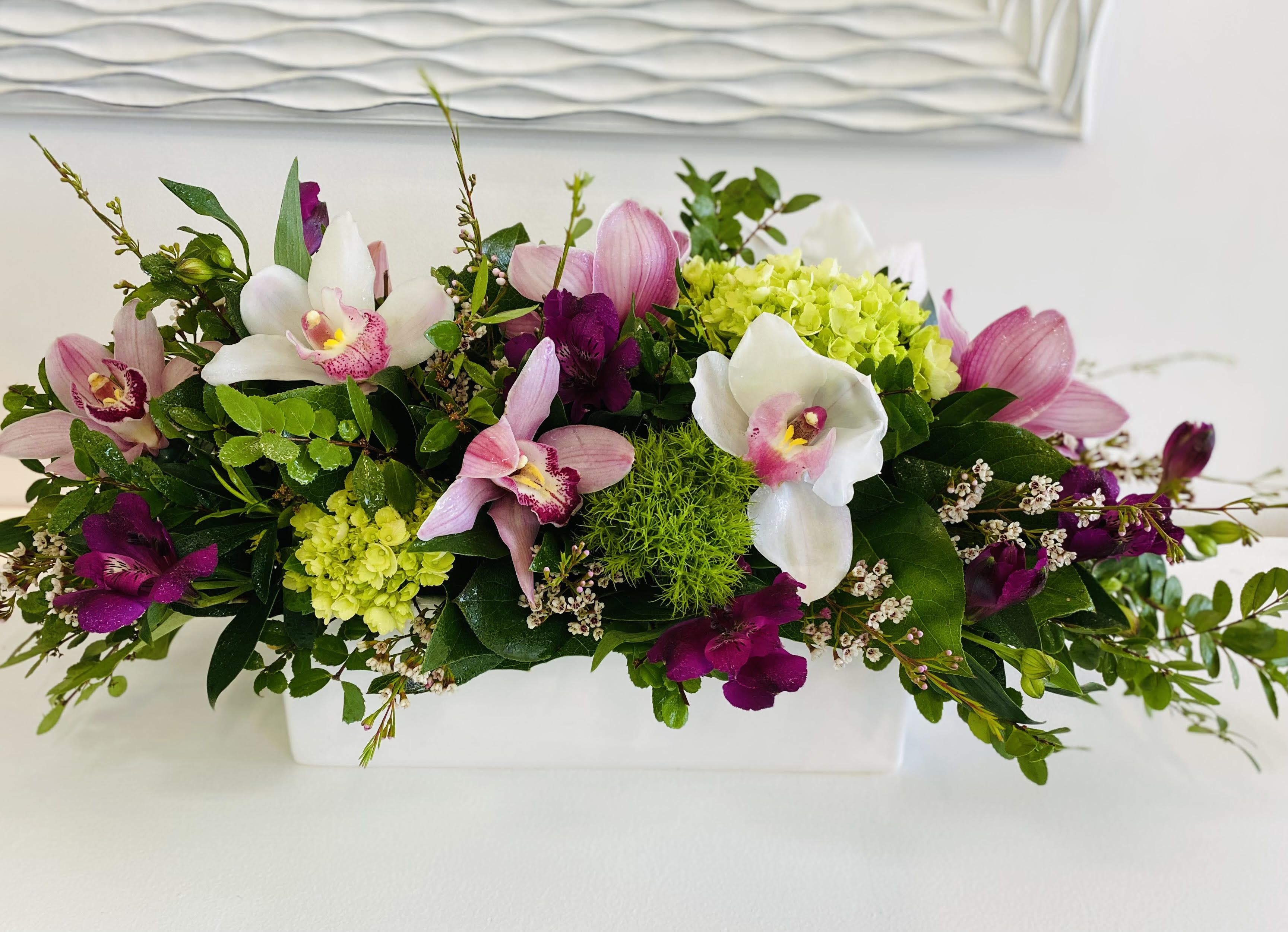Window Box Style Window Box Style - Elegant and exotic, this low and lush arrangement is a stunning blend of textures and color. Featuring delicate cymbidium orchids in shades of blush and white, lime green hydrangea, vibrant dianthus moss, and rich wine-toned blooms, it’s all brought together with fresh greenery and soft accent flowers. Arranged in a sleek white ceramic container, it’s a striking piece perfect for dining tables, coffee tables, or as a thoughtful and refined gift.