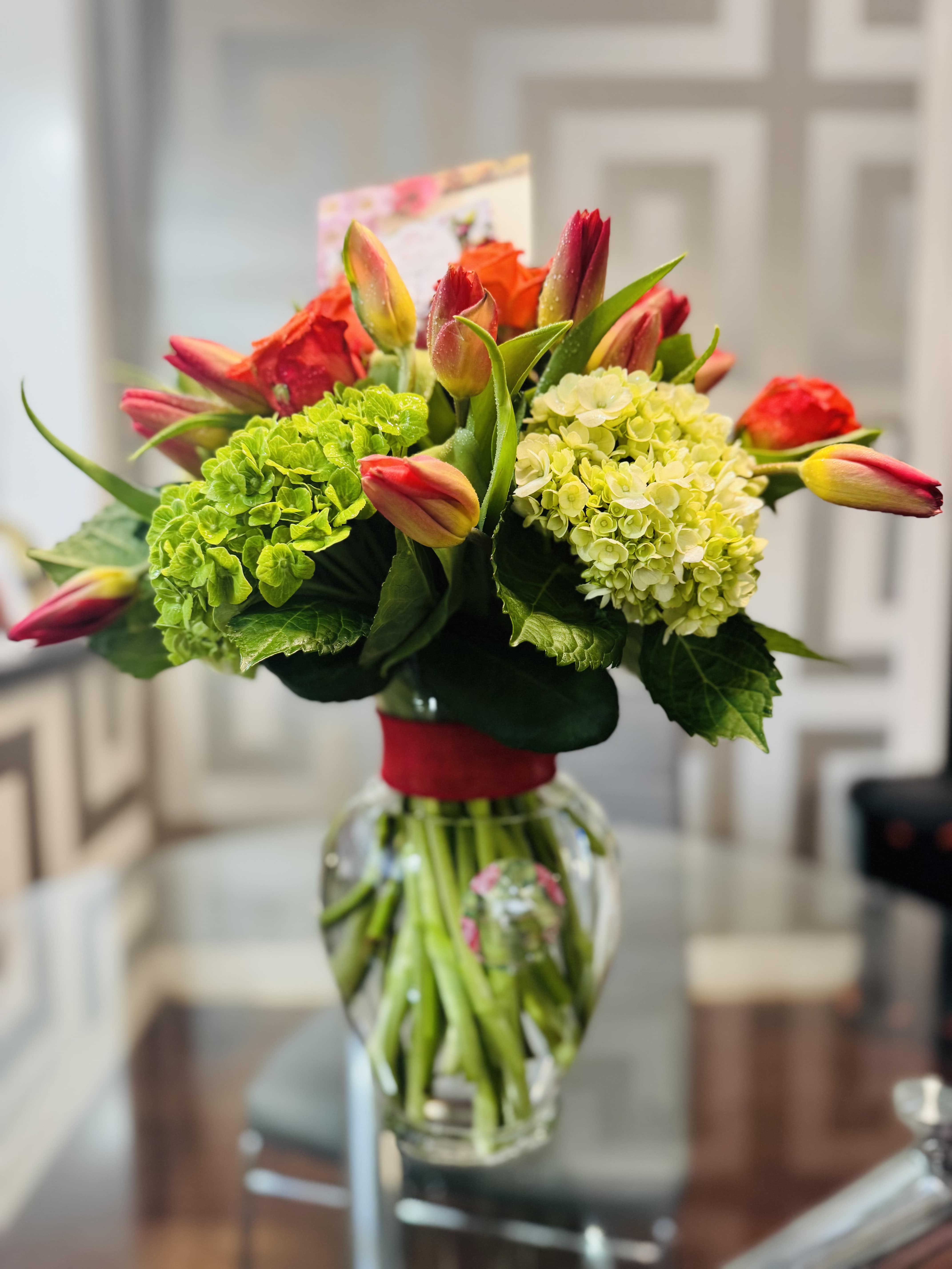 Beautiful Clouds - This bouquet is a vibrant celebration of warmth, energy, and elegance. Bursting with orange tulips and orange roses, it radiates joy, passion, and heartfelt enthusiasm. The Tulips, with their sleek, cup-shaped blooms, bring a sense of fresh beginnings and affectionate emotion. The roses add layers of depth-each velvety petal a note of admiration, desire, and lasting love.  Cradling these vivid blooms are lush green hydrangeas, their voluminous clusters offering a soothing contrast. The green brings balance and harmony, symbolizing renewal, grounding, and abundance.  Together this bouquet feels like a sunlit garden at the height of summer- rich, romantic, and full of life. It's a perfect expression of bold affection, spirited charm, and natural beauty wrapped in golden warmth and earthy elegance.