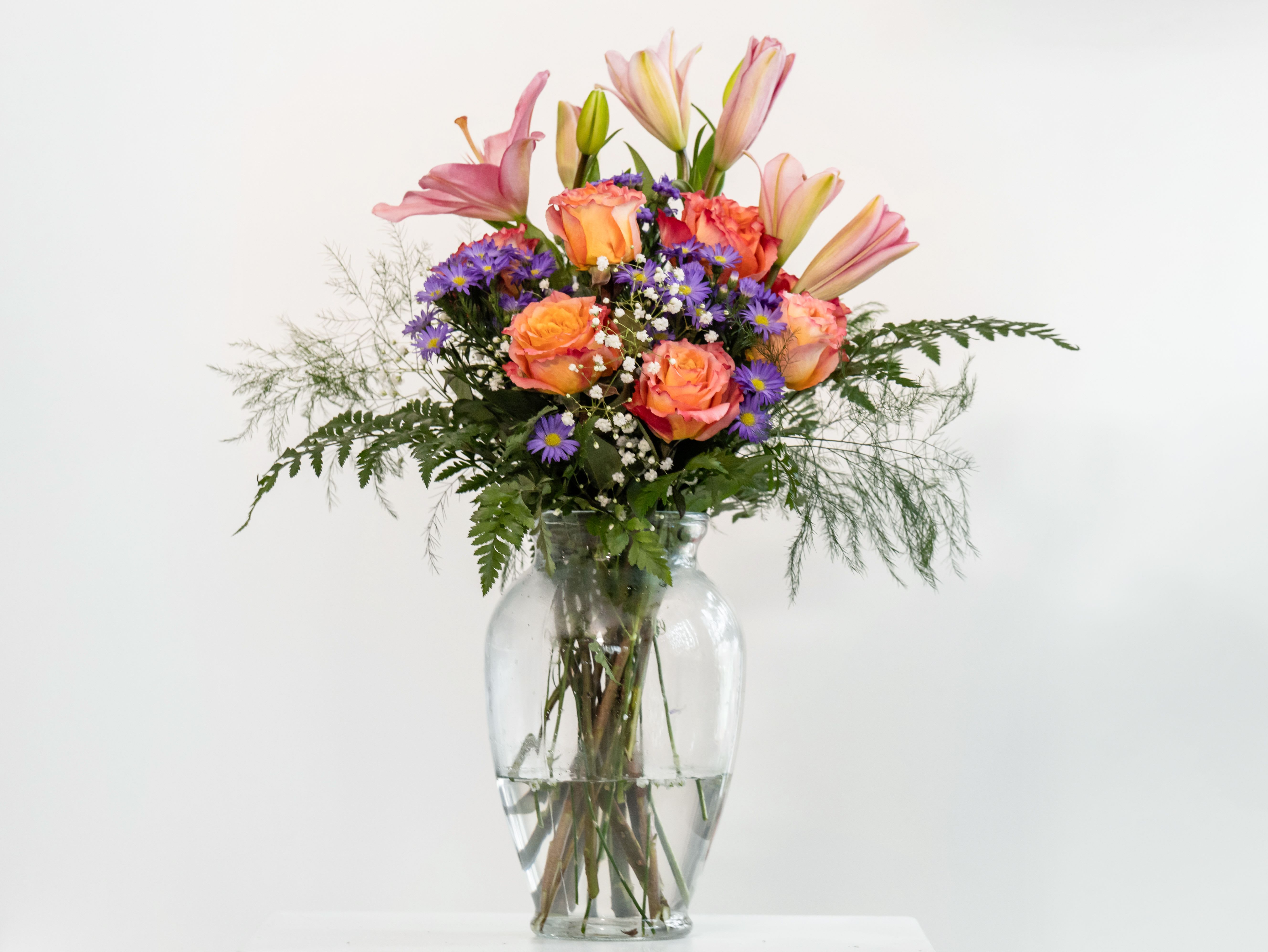 Pretty like your smile by Dixie's Flowers Pretty like your smile by Dixie's Flowers - This vibrant arrangement features a cheerful mix of peachy-orange roses, soft pink lilies, delicate purple asters, and airy baby's breath, all nestled in lush greenery. Designed in a classic clear glass vase, it brings warmth, elegance, and joy to any space—just like a beautiful smile. Perfect for birthdays, anniversaries, or simply brightening someone's day.