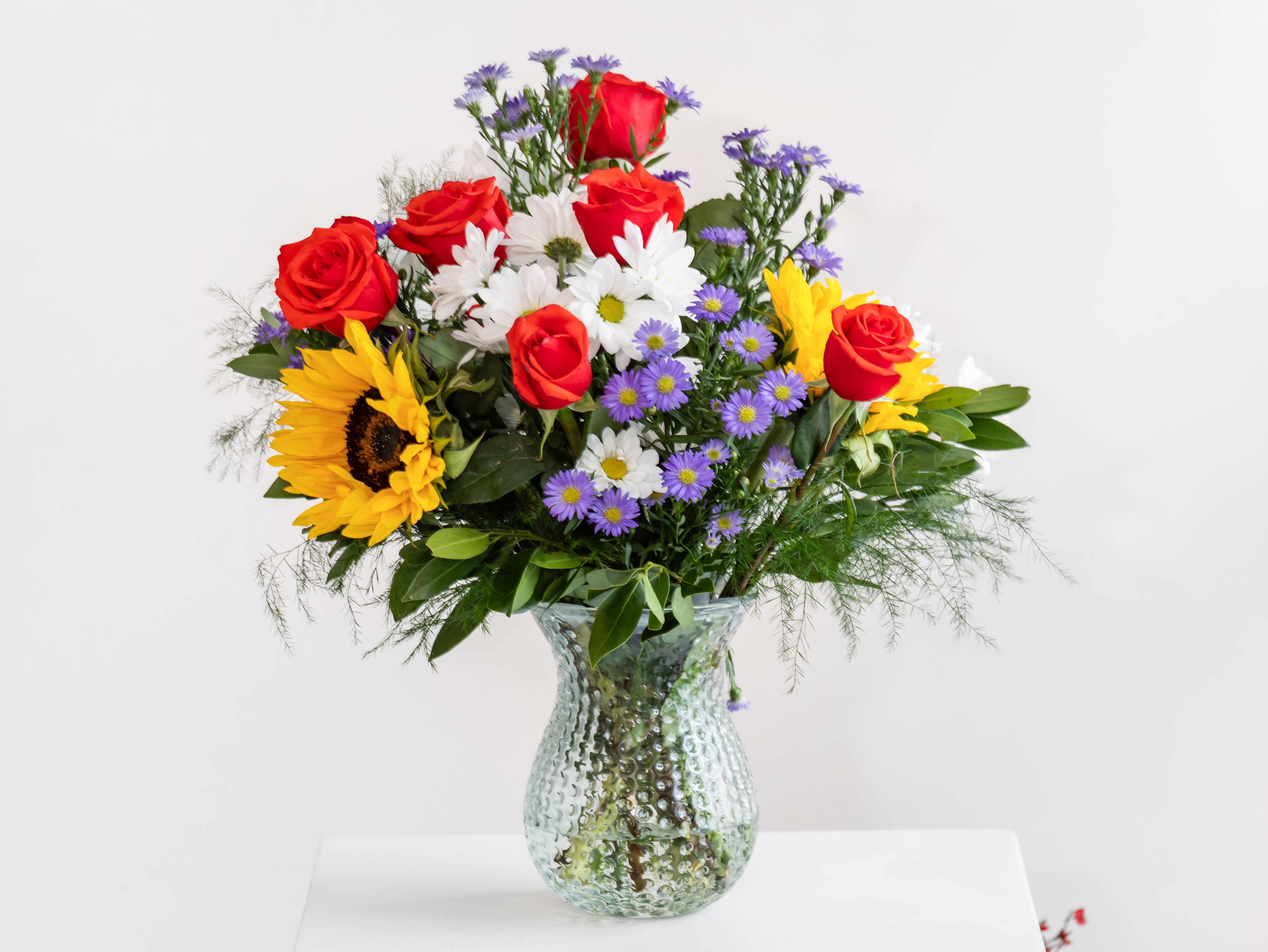 Keeley Boutique by Dixie's Flowers - A joyful burst of color, this lively arrangement features bold red roses, radiant sunflowers, classic white daisies, and charming purple asters. Set in a textured glass vase, it's a celebration of beauty, warmth, and personality—perfect for brightening someone’s day or making any occasion feel extra special.