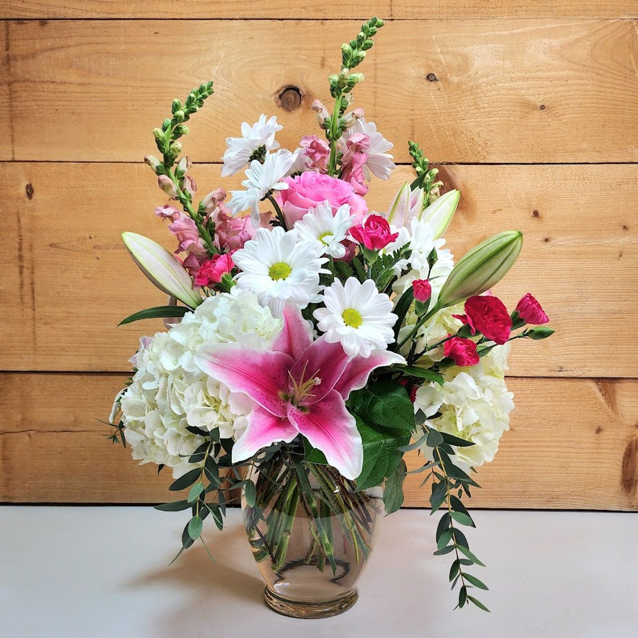 Blooms of Beauty Bouquet - Blooms of Beauty is a captivating floral arrangement that radiates elegance and charm. This stunning bouquet features a harmonious blend of fresh flowers, including vibrant pink roses, delicate white hydrangeas, striking stargazer lilies, cheerful daisies, and lush greenery. Each bloom is thoughtfully arranged in a glass vase, adding a touch of sophistication.  With its soft hues of pink, white, and purple, this arrangement evokes feelings of grace and serenity, making it a perfect gift for any occasion—be it a birthday, anniversary, or a simple gesture of appreciation. The mix of textures and colors creates a balanced yet eye-catching display, making Blooms of Beauty a true statement piece that brightens any space.  Approx 20&quot; H x 14&quot; W