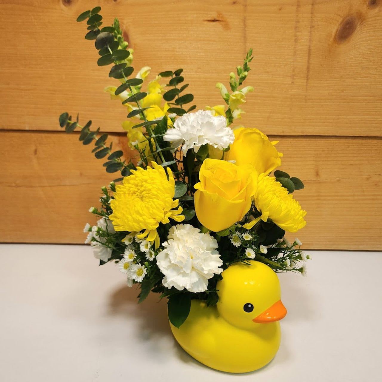 Duckling Delight - Welcome the newest bundle of joy with “Duckling Delight,” a whimsical floral arrangement that’s as charming as it is cheerful. Presented in an adorable ceramic yellow duck container, this design features a sunny mix of blooms that perfectly capture the essence of celebration and happiness.  The arrangement boasts vibrant yellow roses, symbolizing joy and new beginnings, alongside delicate white monte, which adds a touch of elegance and purity. Bright yellow cremones and cheerful yellow snapdragons infuse the design with lively color and texture, while spiral eucalyptus introduces a fresh, green touch that beautifully complements the warm hues.  This enchanting arrangement is ideal for welcoming a baby with a burst of color and a playful nod to the classic duck theme, creating a heartwarming and memorable centerpiece for any baby shower or new arrival celebration.