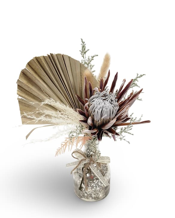 Timeless Token Timeless Token - Timeless Token is a striking dried arrangement featuring a bold protea bloom, fan palm, pampas, and soft textured accents, all artfully gathered in a patterned glass vase with a rustic ribbon tie. A lasting statement piece that brings natural beauty and sculptural form to any space.