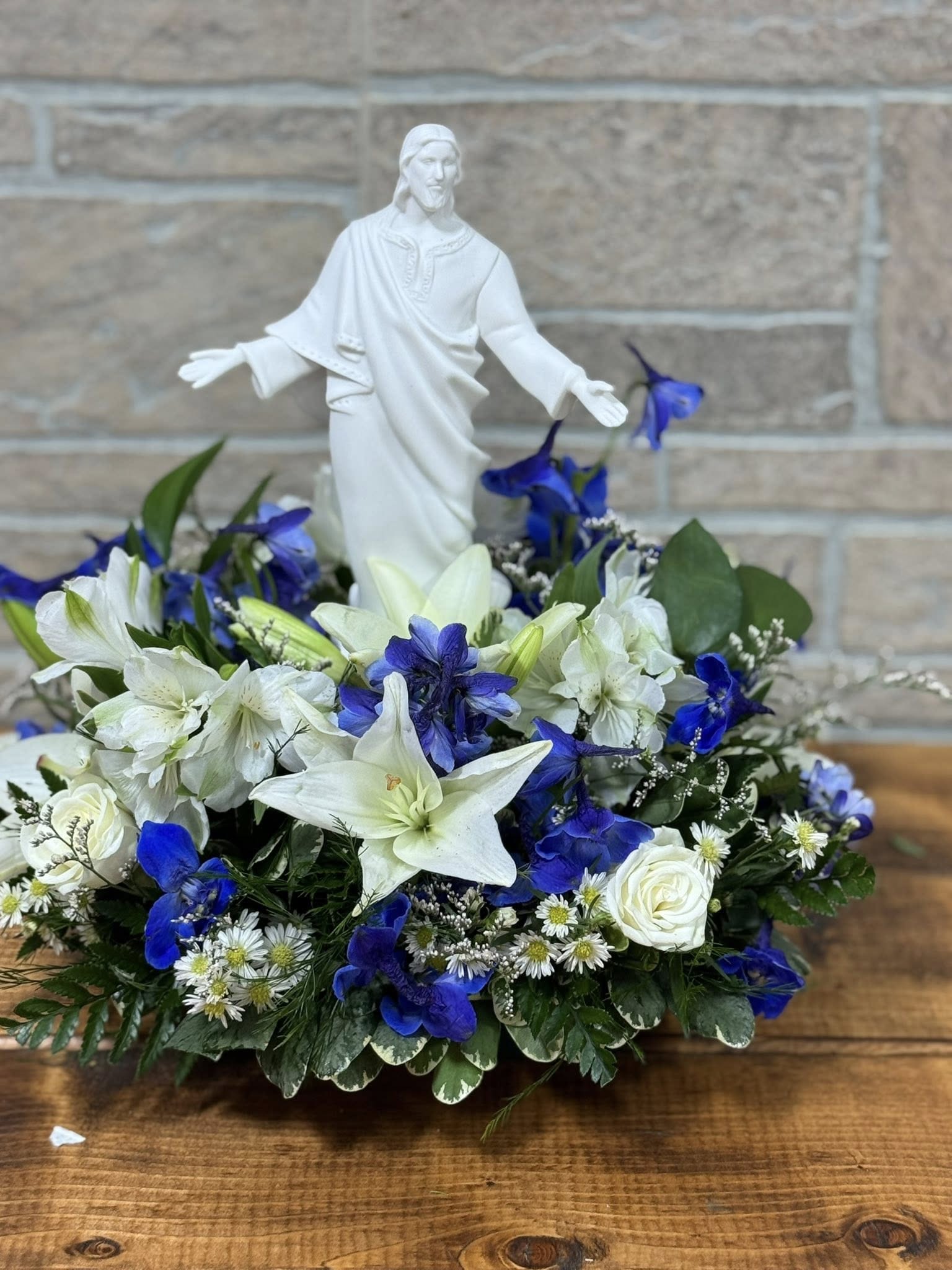 In His Presence - A beautiful expression of faith and comfort, the In His Presence Arrangement centers around a serene ceramic statue of Jesus, surrounded by a lush display of fresh, seasonal blooms. Thoughtfully designed to honor a life lived in faith, this arrangement offers a peaceful presence and a reminder of eternal love and hope. Ideal for funeral services, memorials, or as a lasting tribute in a place of remembrance, it brings solace and spiritual reflection to those who grieve, while celebrating a soul now resting in His presence.