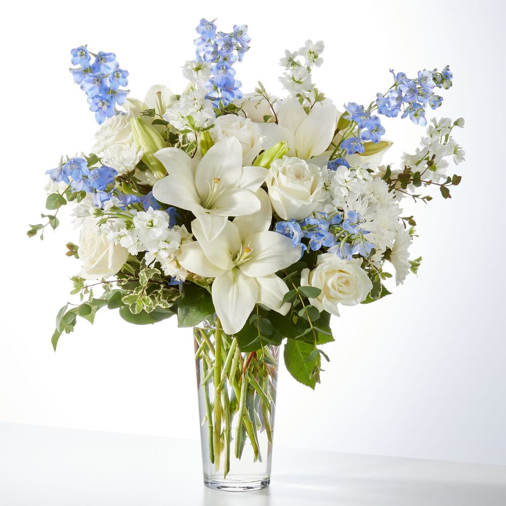 White Lilies and Blue Delphiniums  - Bring a sense of peace and elegance to any moment with this exquisite white and blue flower arrangement from Miami Flowers Design, the leading florist in Miami, Florida. This graceful bouquet features a soothing mix of white lilies, ivory roses, white chrysanthemums, and delphinium in soft sky blue. Accented with fresh greenery and delicate fillers, the design is presented in a tall, clear glass vase to highlight each stem’s natural beauty.  Perfect for expressing sympathy, celebrating new beginnings, or simply adding a touch of serenity to your space, this bouquet is a timeless choice for all occasions. Handcrafted by expert floral designers at Miami Flowers Design, each arrangement is created with precision and care to ensure freshness and artistic presentation.  Offering same-day flower delivery in Miami, Coral Gables, Brickell, and surrounding areas, we specialize in bringing elegance and comfort to your doorstep. Whether you’re shopping for a thoughtful gift or a calming centerpiece, this arrangement is ideal for both home décor and heartfelt gestures.  Trust Miami Flowers Design for premium floral arrangements that capture emotion and style. Shop online today and discover why we’re a top-rated florist in Miami for beautiful, meaningful flowers delivered with care.