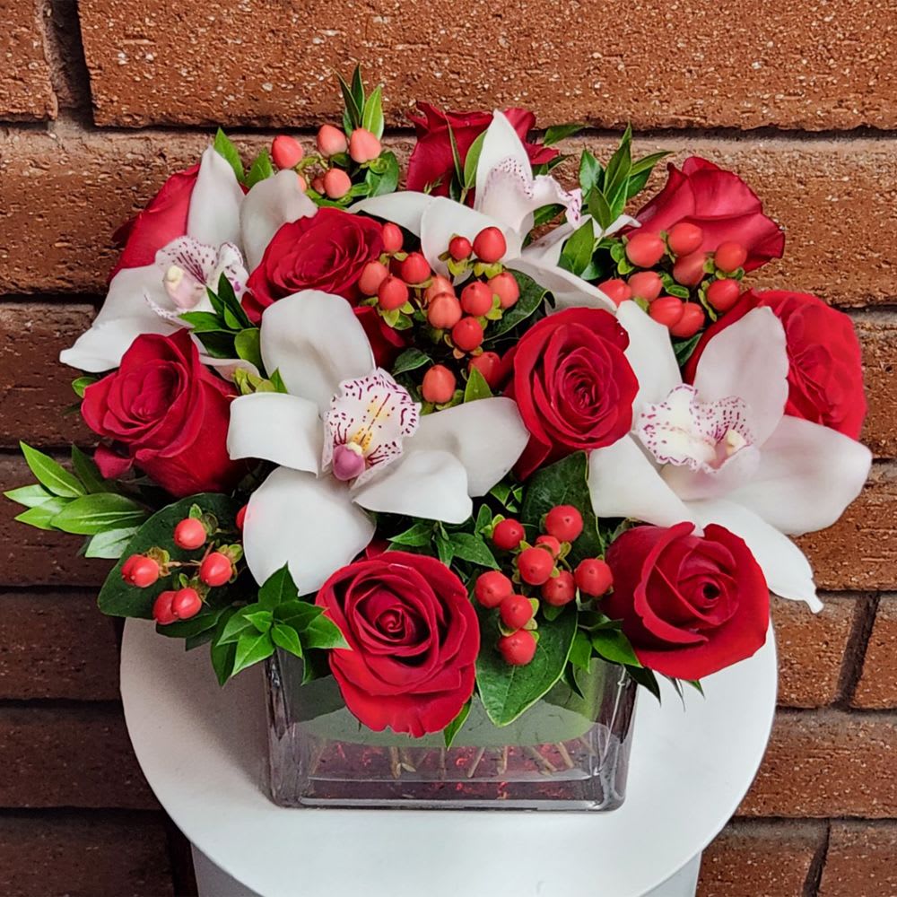Striking Red Roses and White Orchid Cube - A striking fresh cut floral arrangement of premium red roses, white cymbidium orchid blooms, red berries and fresh garden greens. Standard size is approximately 8in (W) x 8in (H). Deluxe version is larger and features more premium blooms along with a jumbo glass cube vase.  Standard (Stem Count) – 9 Roses, 4 Orchid Blooms, 5 Red Berries &amp; Fresh Garden Greens - 6in Cube Vase  Deluxe (Stem Count) – 18 Roses, 7 Orchid Blooms, 8 Red Berries &amp; Fresh Garden Greens - 8in Cube Vase  Care Tips: Place your bouquet in a cool location. Don't put the arrangement in direct sunlight, near heating or cooling vents, in drafty places, directly under ceiling fans, or on top of televisions or radiators. Check water level daily, keep the vase full with clean water. Change water every 2-3 days and apply a sharp fresh cut to the stems. This process will ensure extended flower's life span.