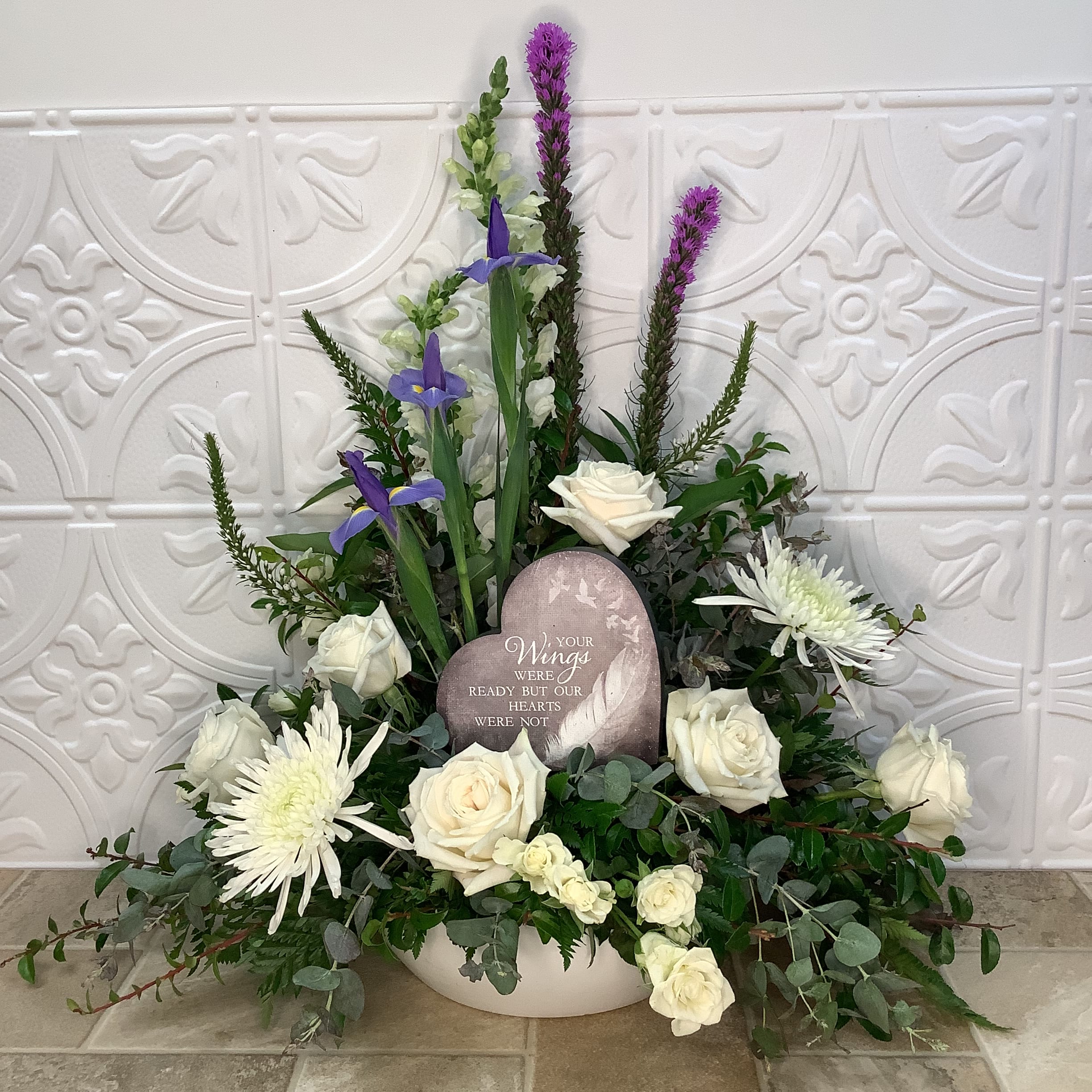 Wings Wings - This large floral arrangement, in a white, shallow bowl, is ideal for a sympathy or memorial occasion, with the centrally placed, heart-shaped plaque that reads "Your wings were ready but our hearts were not." Large white spider mums, white spray roses, and white snapdragons offer a calm and peaceful look. Touches of color, texture and height are added by accents of veronica, purple liatris and blue iris. Foliage in different green hues adds depth. Colors cn be changed upon request. Approximate size: 28" wide x 26" high. Primary flowers: chrysanthemum, rose, snapdragon.