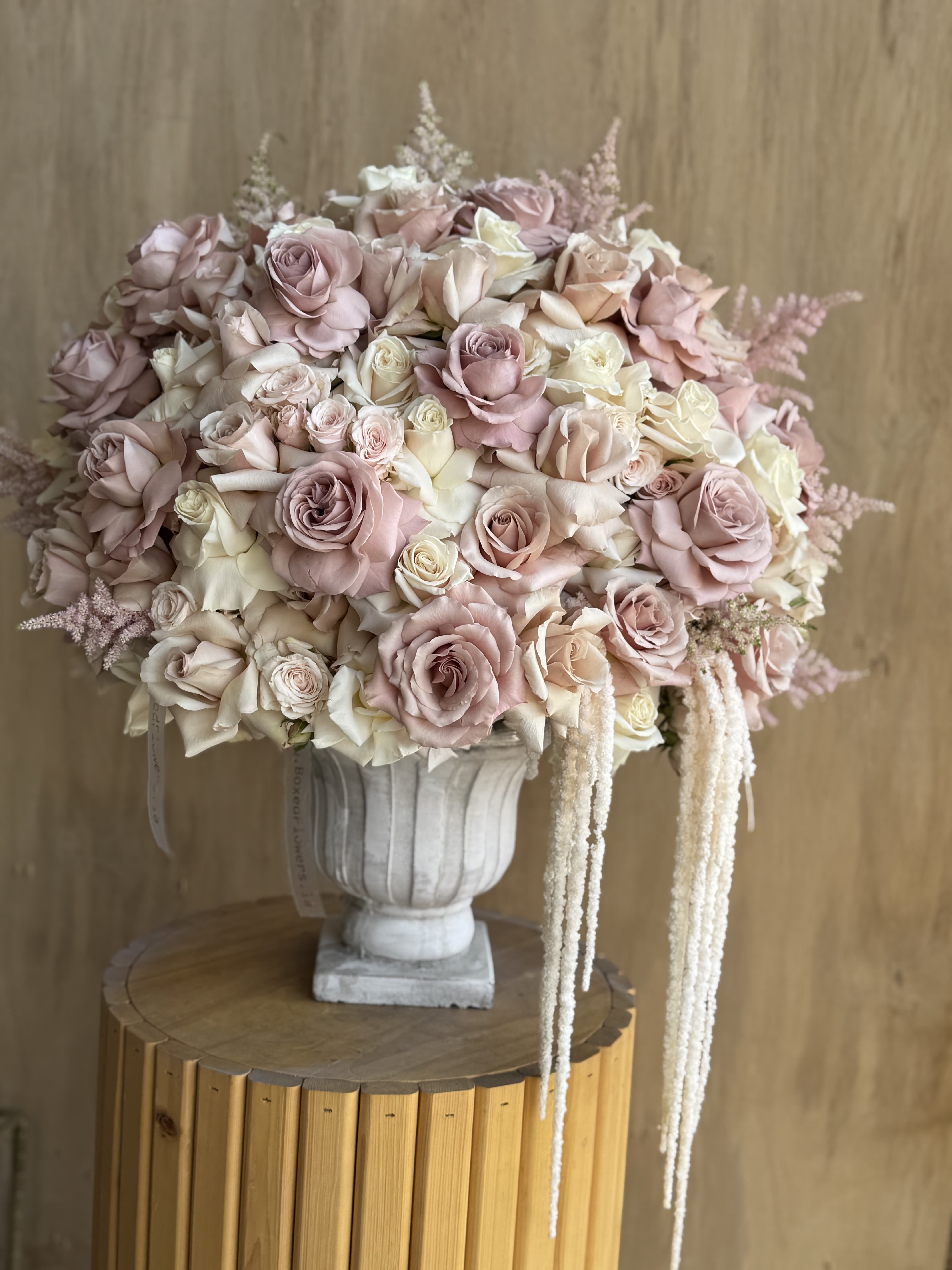 Blush Embrace - An exquisite dome of soft romance, Blush Embrace is a refined composition of pale pink, cream, and ivory roses artfully arranged in a classic stone urn. Accented with soft, feathery astilbe and cascading amaranthus, this arrangement exudes timeless elegance and femininity. Perfect for weddings, anniversaries, or luxurious home décor, it offers a poetic expression of grace, love, and delicate beauty.