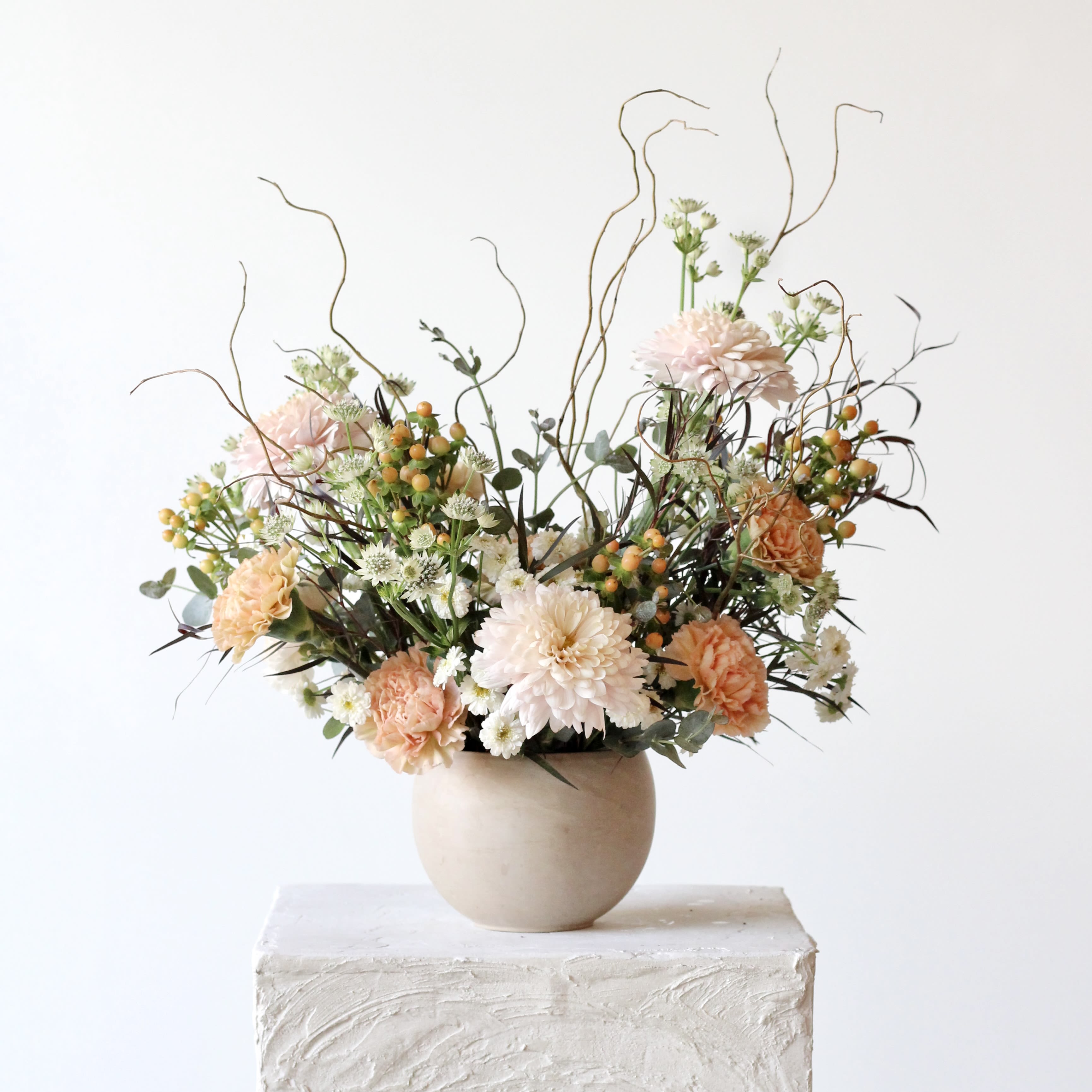 Very Demure - Our &quot;Very Demure&quot; arrangement offers a refreshing alternative to traditional florals. Featuring soft pink cremone mums, elegant lege marrone carnations, delicate peach hypericum, and artful curly willow, this arrangement brings a subtle yet distinctive color palette to your table. Arranged in the Astrid pot—a tan round container crafted from natural clay—it's the perfect choice for those looking to celebrate in their own unique way. 