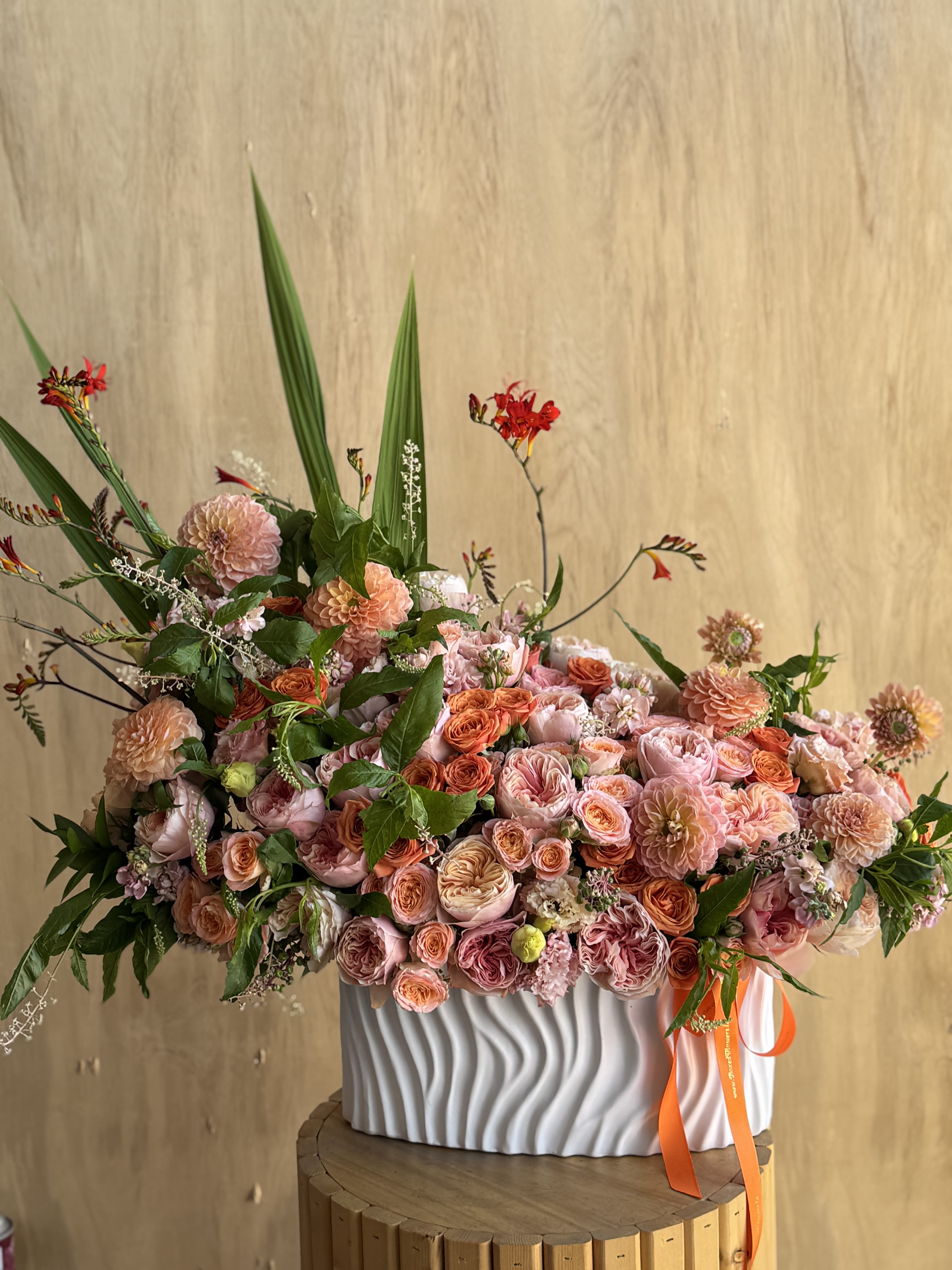 Tropical Sunset - Bursting with the vibrant energy of a tropical sunset, that is a joyful arrangement featuring a rich blend of coral, peach, and apricot garden roses, dahlias, and spray roses, accented by vivid greenery and striking red crocosmia. Designed in a modern white ceramic vase with sculpted waves and finished with a bold orange ribbon, this piece radiates warmth, creativity, and celebration. Perfect for grand openings, birthdays, or summer events, it’s a visual symphony of color and style.