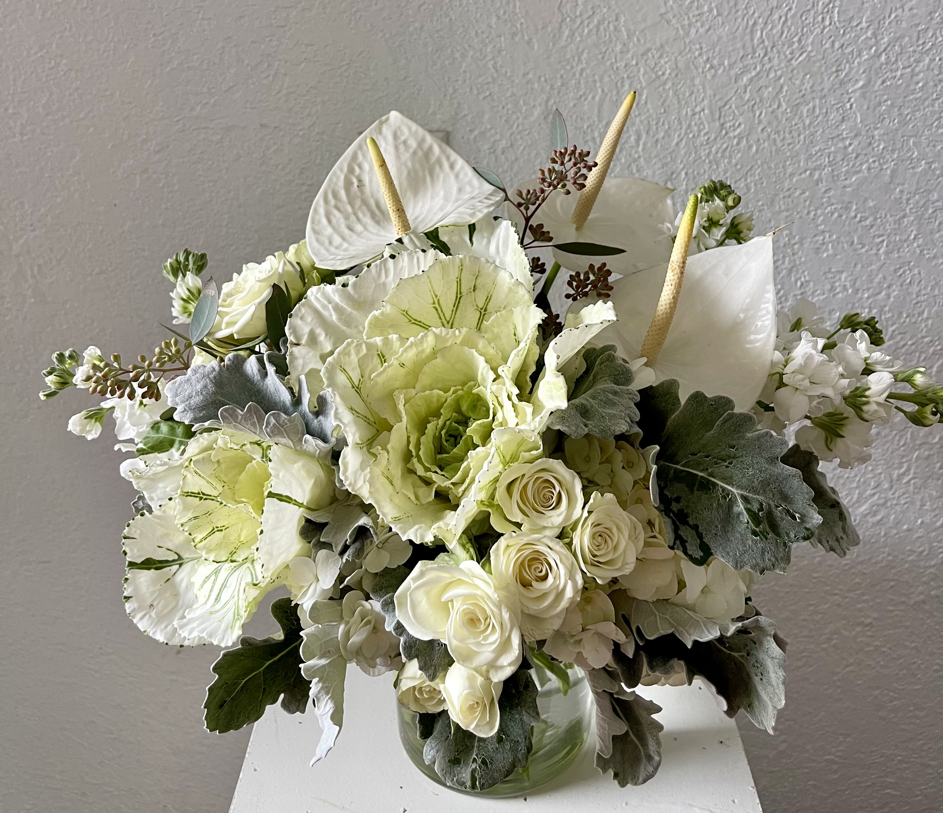 Frosted Elegance - Chic and sophisticated, Frosted Elegance is a striking all-white arrangement that blends modern style with soft, natural texture. Featuring sculptural white anthurium, delicate spray roses, fragrant stock, hydrangeas, ruffled kale, dusty miller, and seeded eucalyptus, this design brings a cool, refined presence to any setting.  Expertly arranged by Julia’s Floral and available for same-day flower delivery in Dallas, Frosted Elegance makes a graceful statement for sympathy, celebration, or everyday luxury