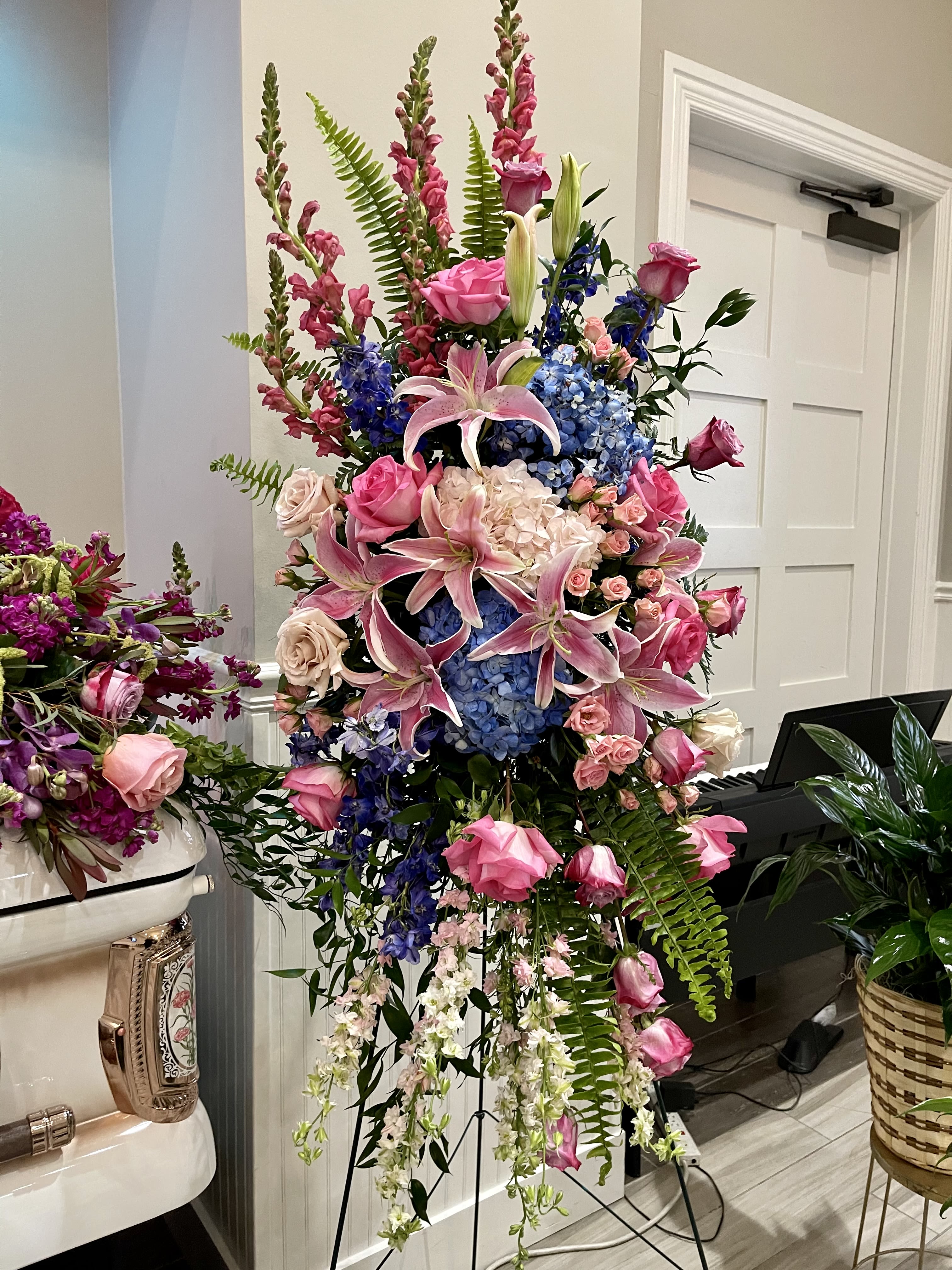 Blooming Legacy  - A lush and heartfelt tribute, Blooming Legacy honors a vibrant life with a graceful display of jewel-toned blooms. This standing spray features blue hydrangeas, elegant roses, larkspur, snapdragons, blue delphinium, and spray roses, complemented by sword fern, ruscus, and mixed greenery. The arrangement’s vertical shape and vivid colors create a striking presence, symbolizing strength, beauty, and lasting memory.  Expertly designed by Julia’s Floral and available by advance order for delivery in the Dallas area, Blooming Legacy pairs beautifully with our Timeless Treasure casket spray or stands powerfully on its own.