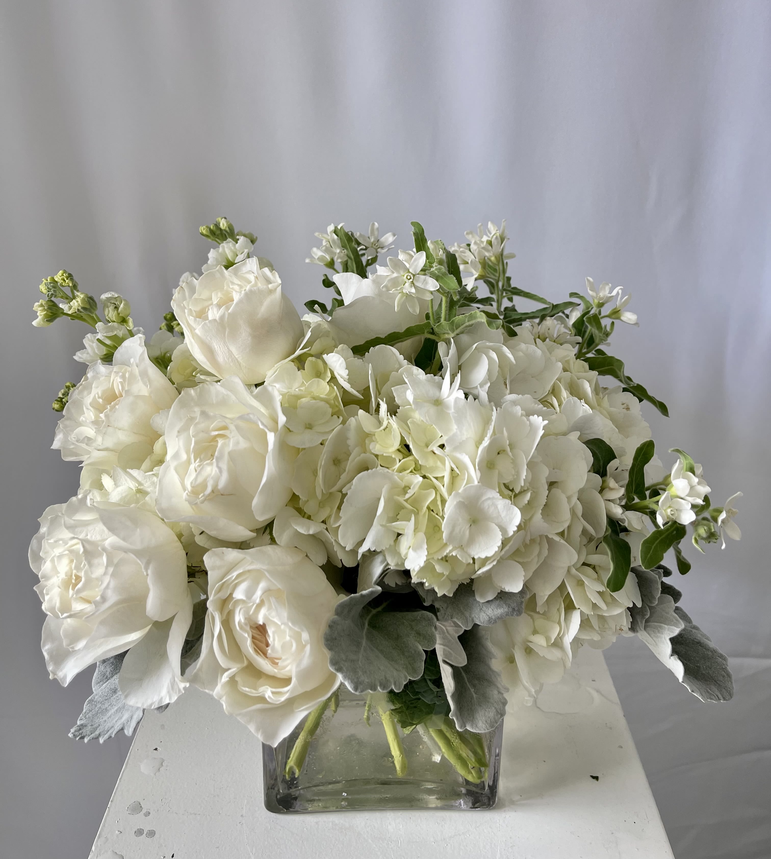 Pure &amp; Simple - Elegant in its understatement, Pure &amp; Simple is an all-white arrangement that speaks volumes with its soft textures and timeless beauty. This serene design features lush white hydrangeas, classic roses, delicate tweedia, and fragrant stock, all accented with silvery dusty miller for a soft, refined touch.  Handcrafted with care by Julia’s Floral, this arrangement is available for same-day flower delivery in Dallas — perfect for expressing sympathy, gratitude, or simply bringing calm sophistication into any space.