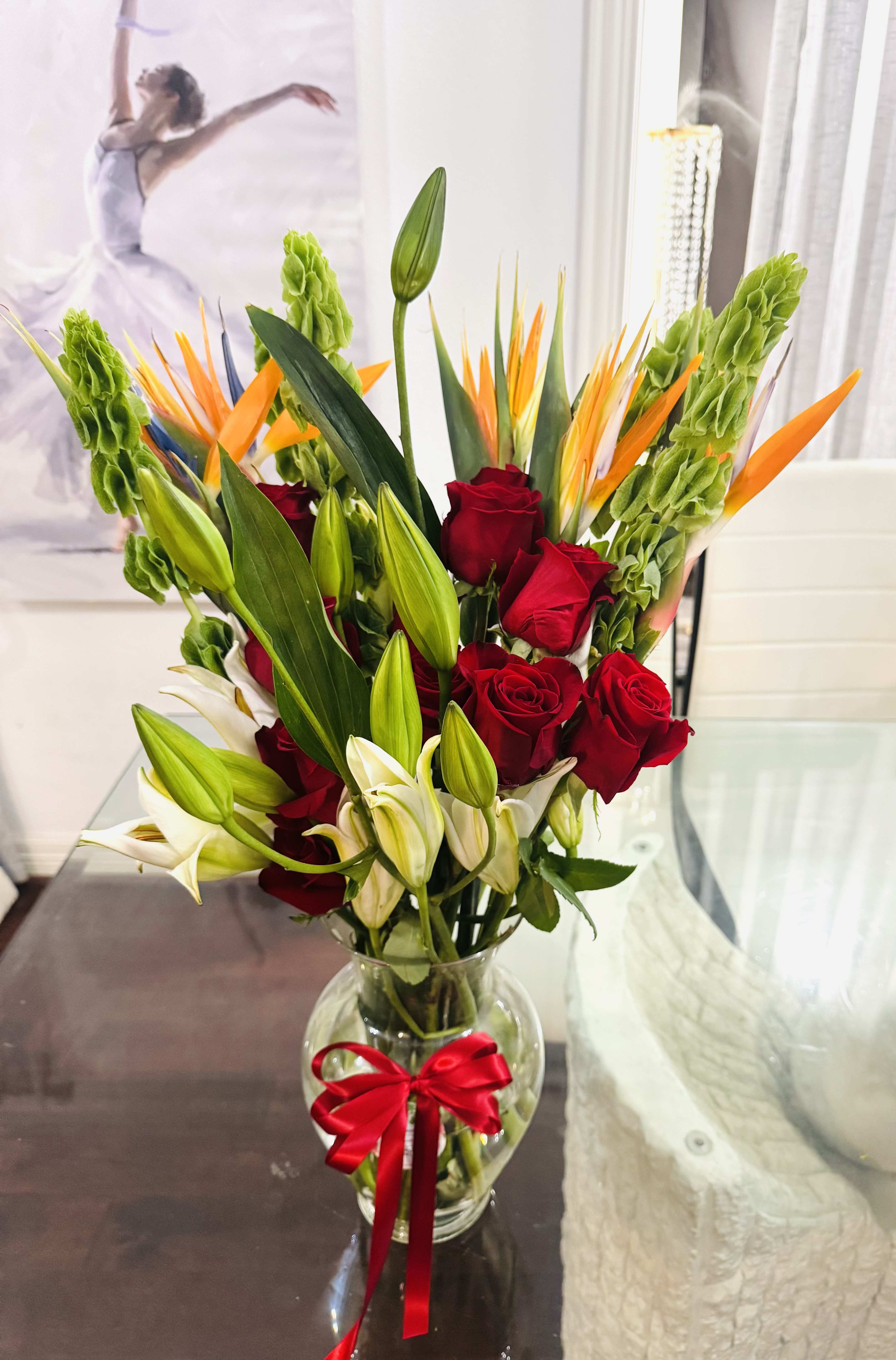 Beautiful Bouquet - A striking and exotic arrangement, this bouquet bursts with passion and vitality. Bold red roses stand at the heart, their velvety petals rich with romance and intensity. Surrounding them, with lilies gracefully, their trumped-shaped blooms adding softness and sophistication. Rising boldly above, the Birds of Paradise lend an architectural, tropical flair- their vivid orange, evoking a sense of freedom and adventure. 